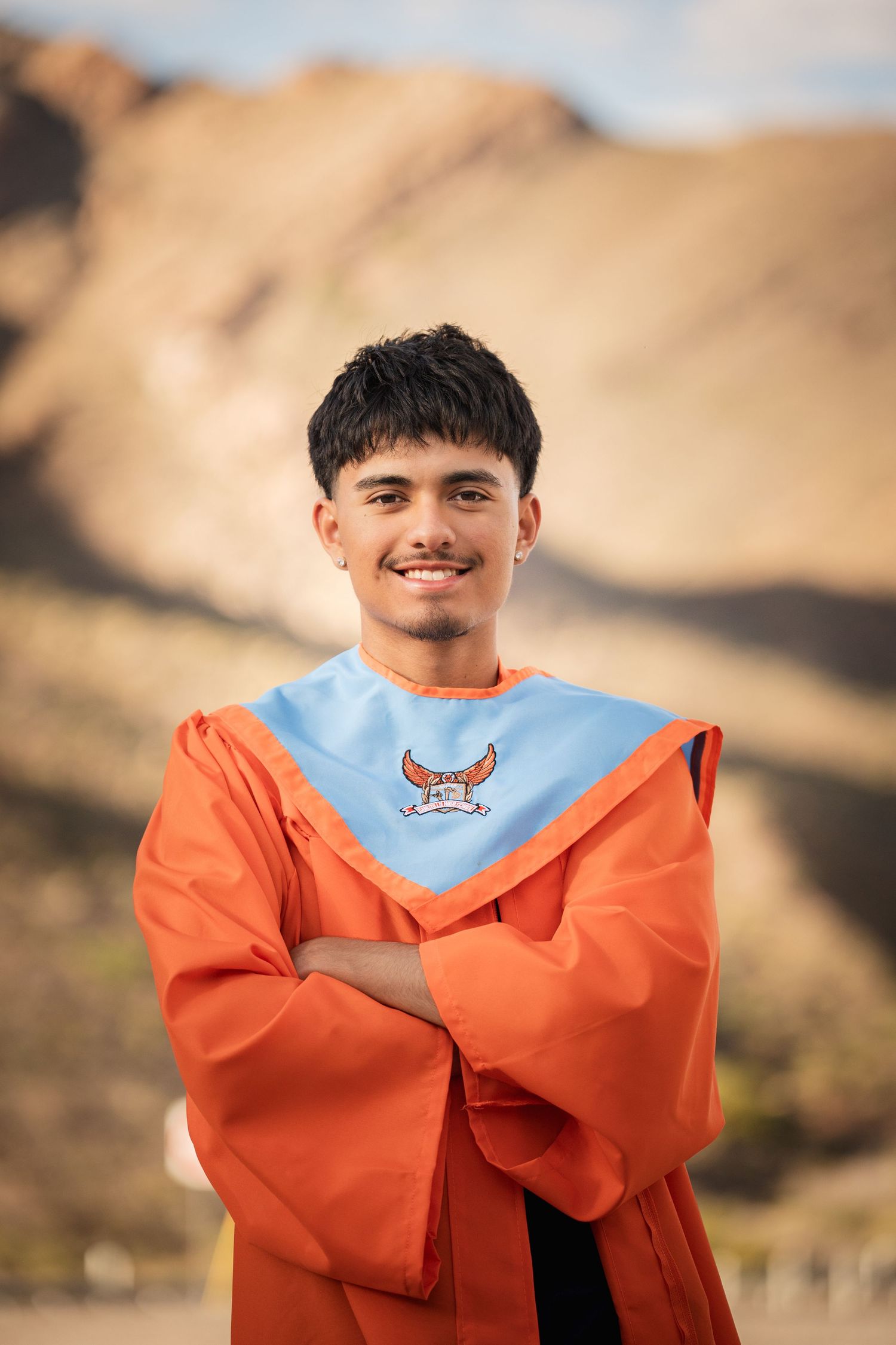 Smiling young man in orange graduation gown with blue collar stands arms crossed before desert hills.