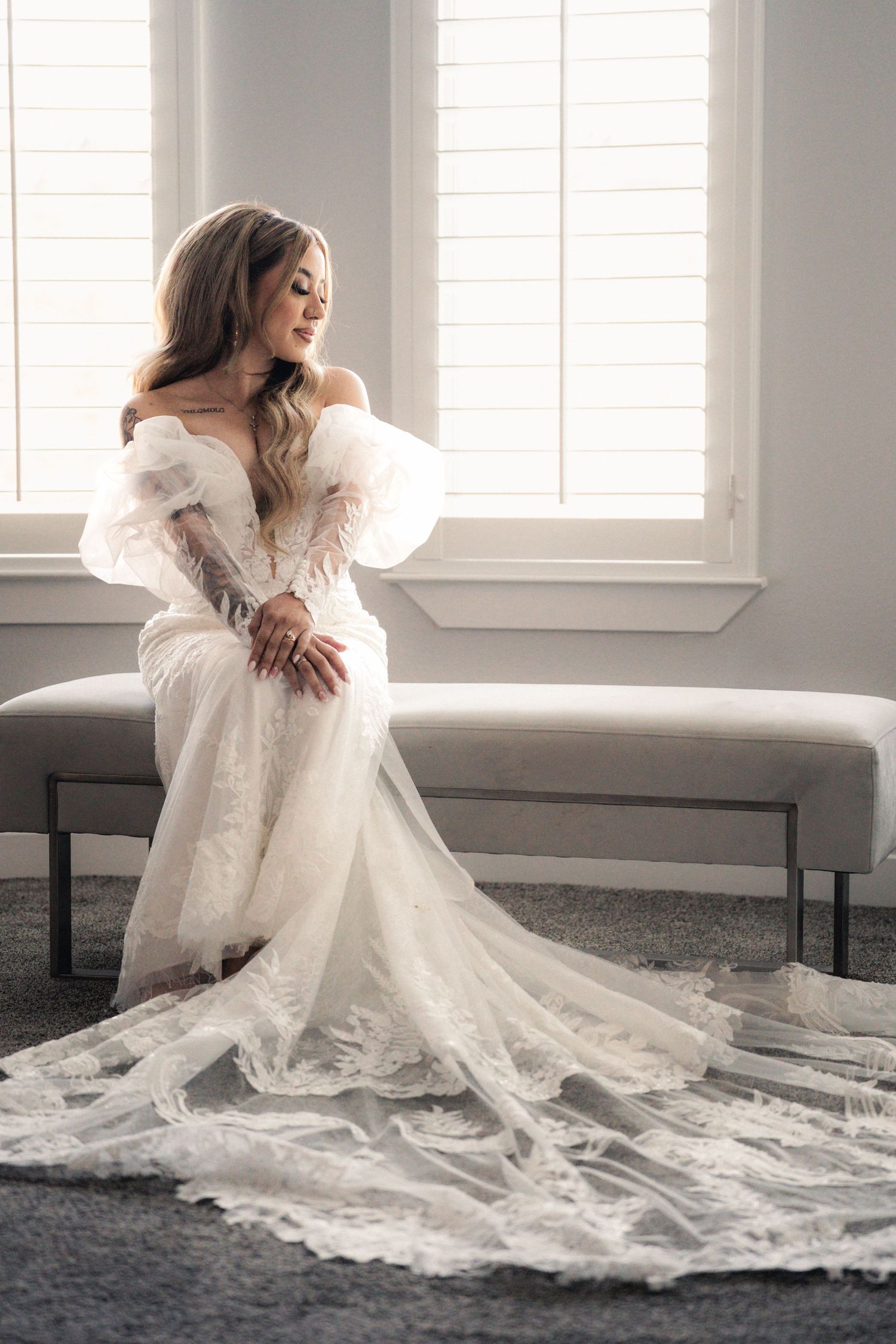 A bride in a flowing white lace gown sits gracefully on a light-colored sofa in a bright, airy room.