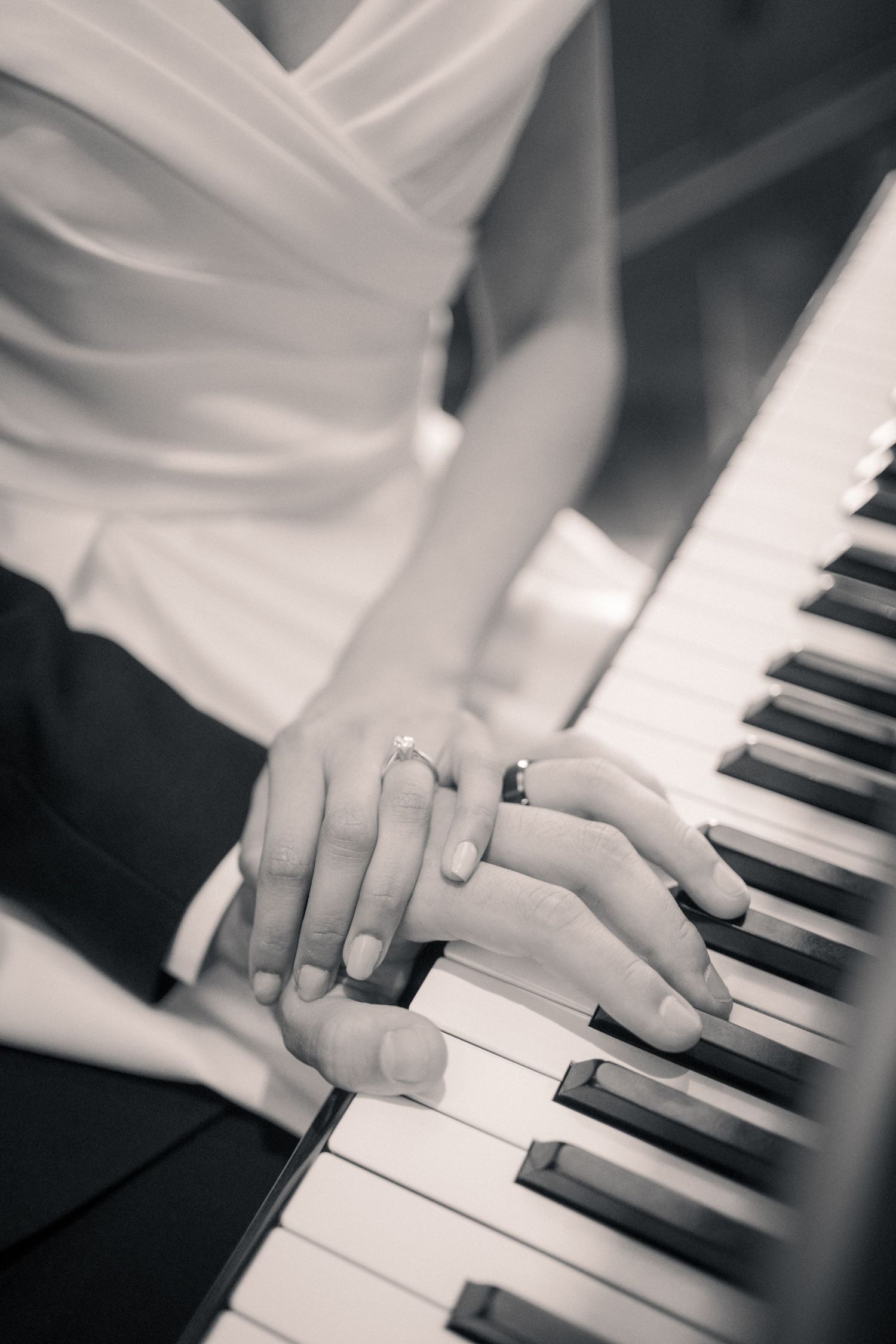 Close-up of hands with wedding rings resting on piano keys in black and white.