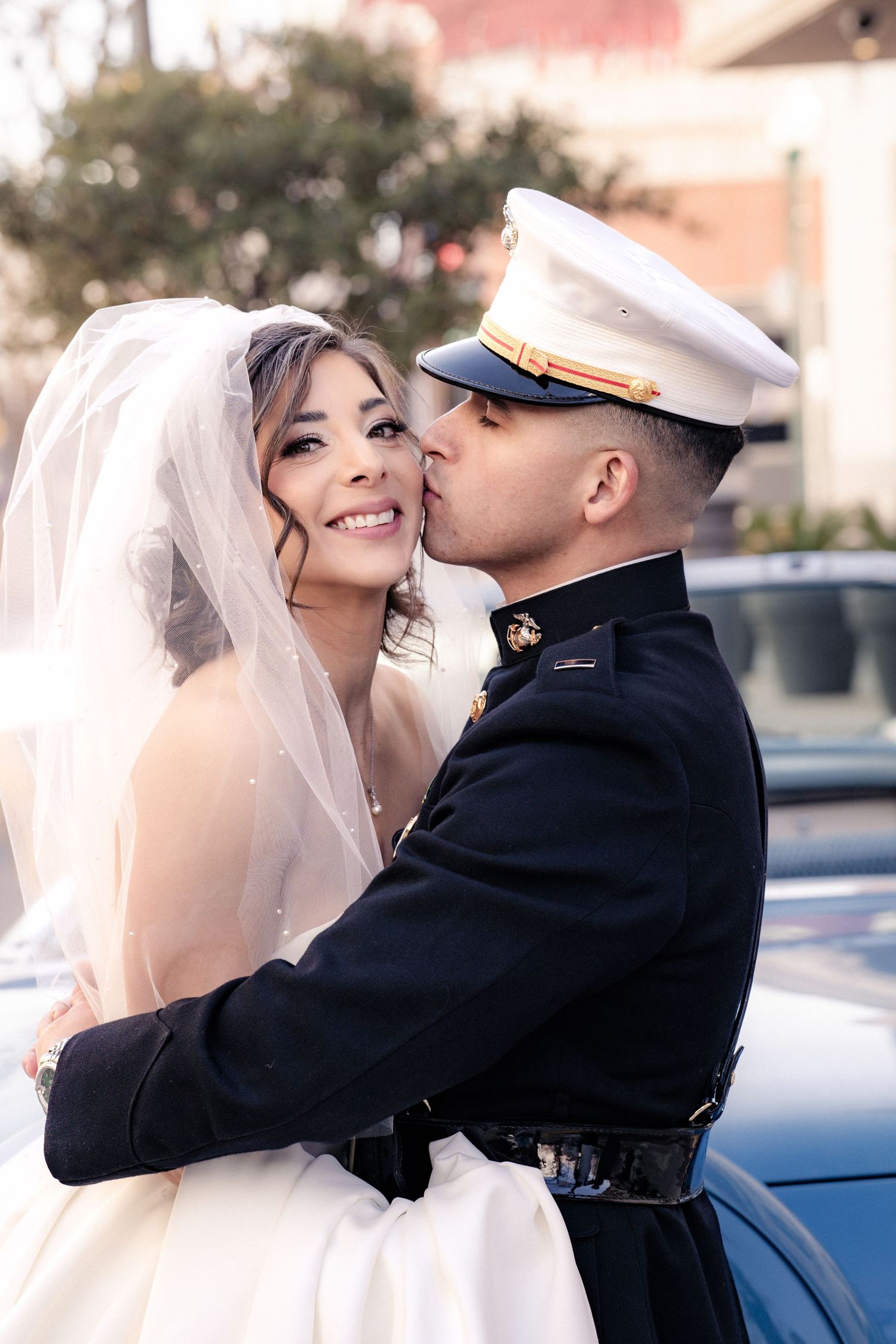 Military groom in dress uniform kissing bride in white wedding gown and veil outdoors near a classic car.