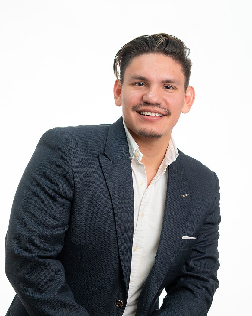Professional in a dark suit jacket and white shirt poses while leaning forward against white background.