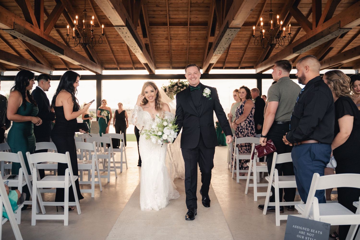 Wedding ceremony guests seated in white chairs under a rustic wooden beam ceiling in an indoor venue.