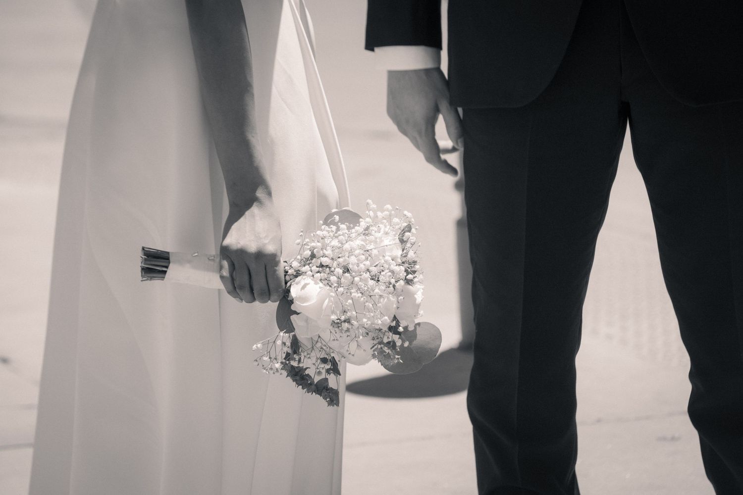 A close-up black and white photo of a wedding couple holding hands and a bridal bouquet.