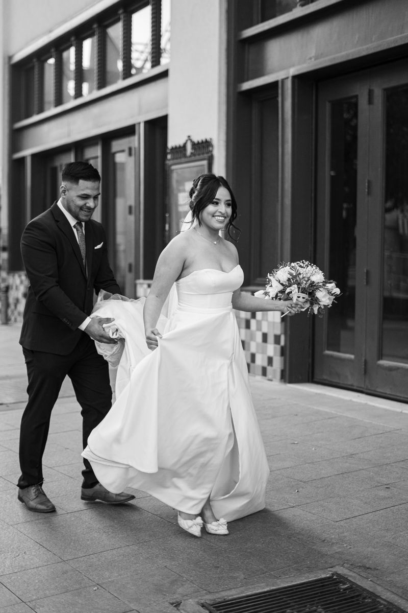 A couple walk together outdoors, with the bride in a flowing white dress and groom helping with the train.