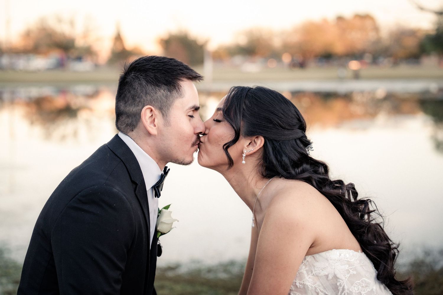 A bride and groom share a romantic kiss outdoors near a reflective pond at golden hour during their wedding.
