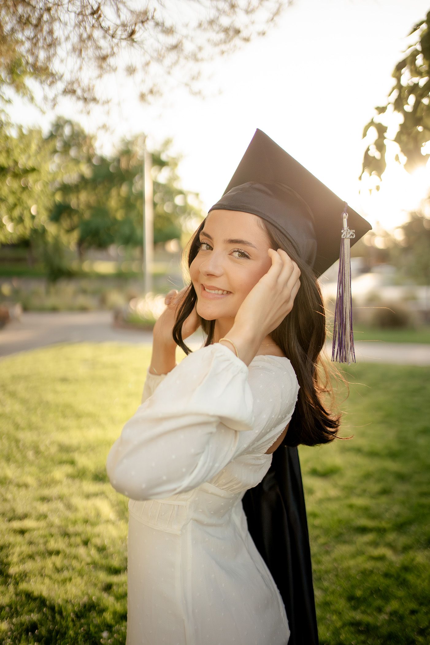 Graduate in white sweater and cap poses joyfully in sunlit grassy field during golden hour celebration photoshoot.
