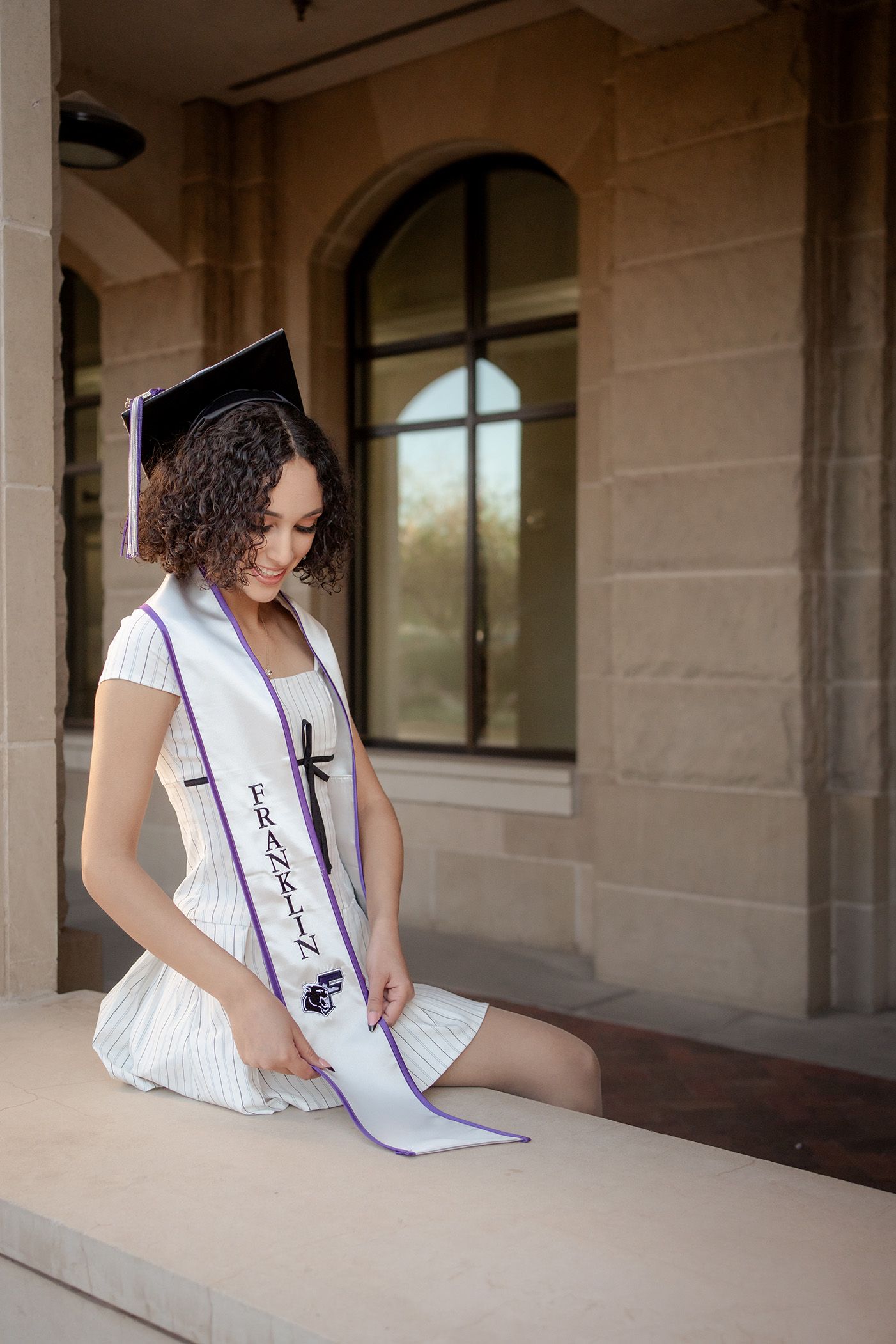 A graduate in a white dress and graduation sash sits thoughtfully on stone steps of a building with arched windows.