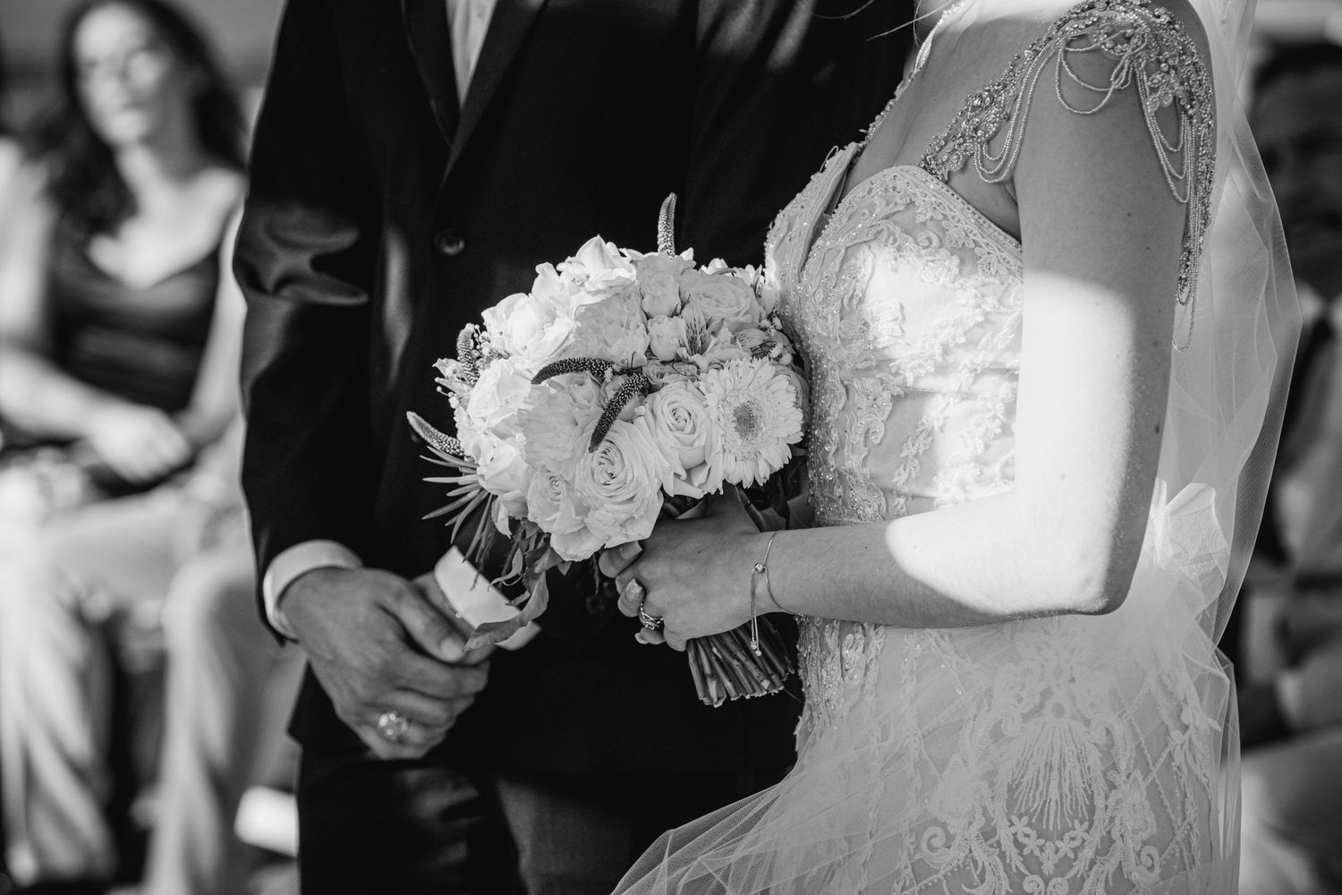 Black and white wedding photo of bride holding a floral bouquet while standing beside groom during ceremony.