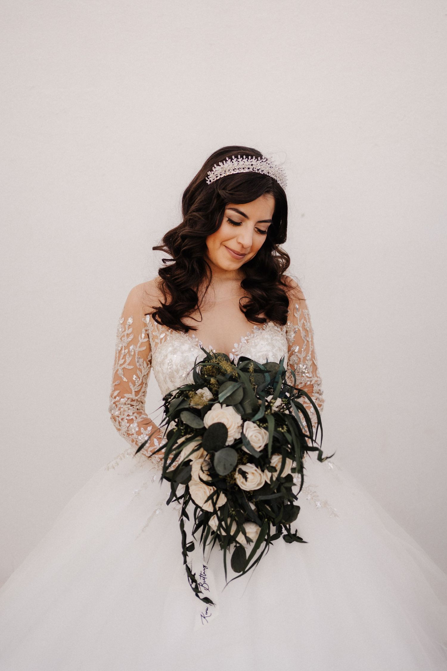 Bride in white lace ball gown with tiara holding cascading bouquet of white roses and greenery.