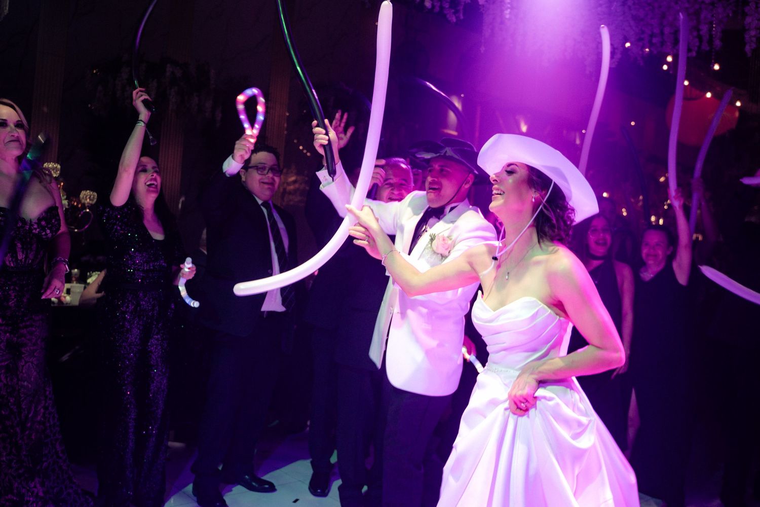 Bride and groom celebrate at wedding reception with guests waving balloon wands under purple lights.