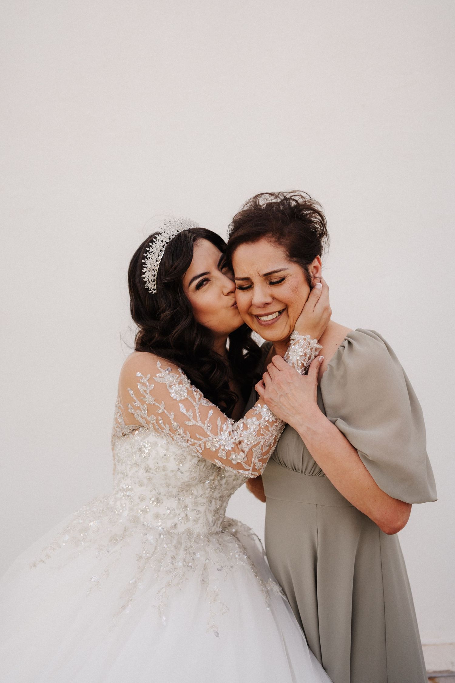 A bride in a white lace gown and tiara kisses a smiling woman in a sage dress on the cheek.