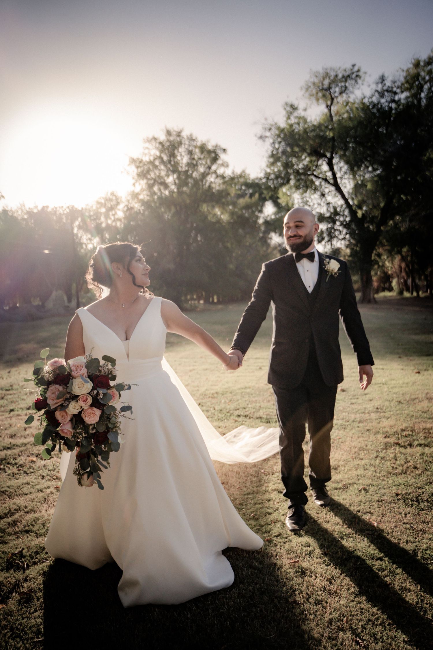 Newlyweds hold hands while walking at sunset in a dreamy outdoor setting.
