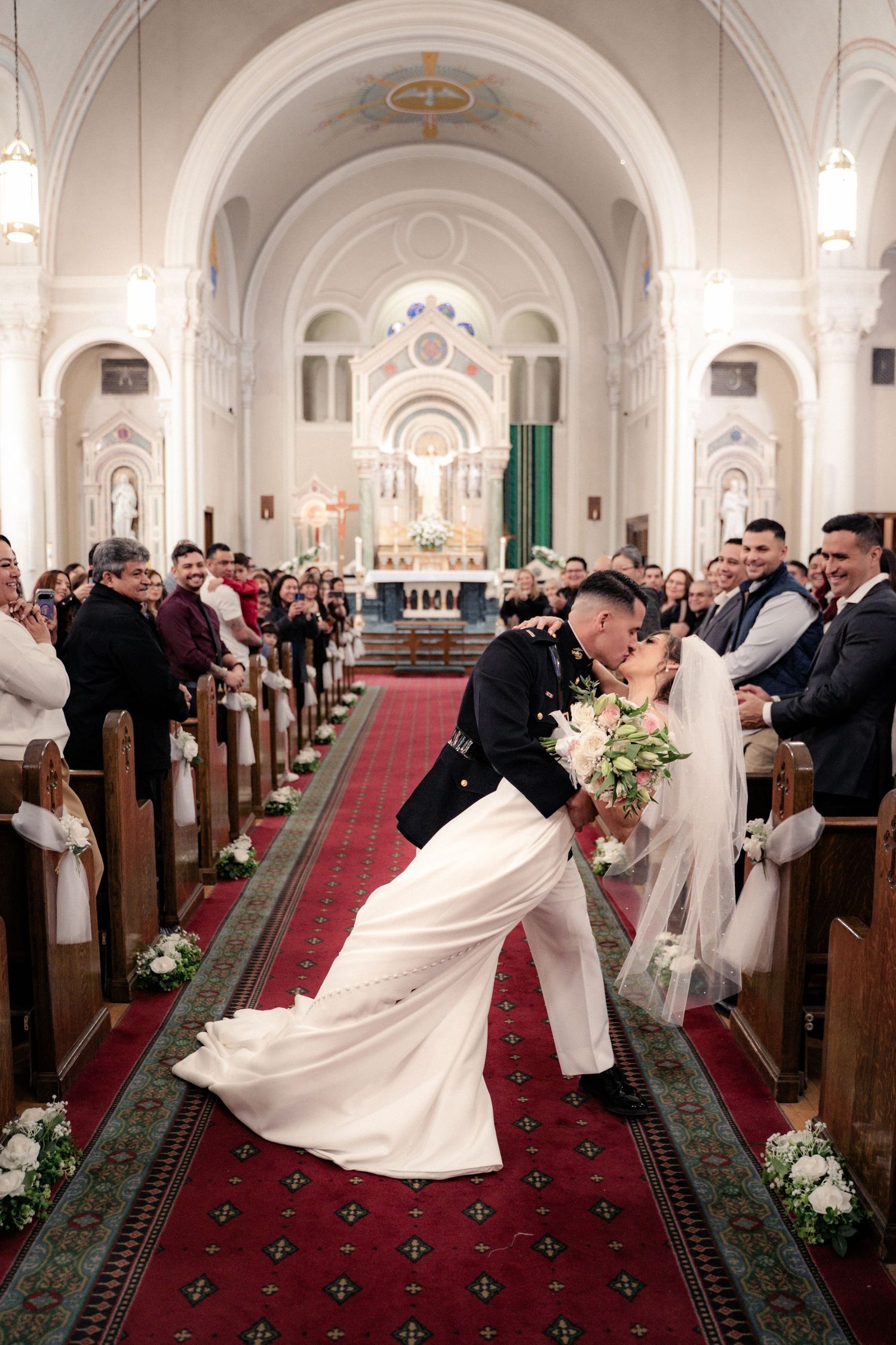 Bride and groom share a romantic dip kiss in a stunning white church aisle during their wedding ceremony.