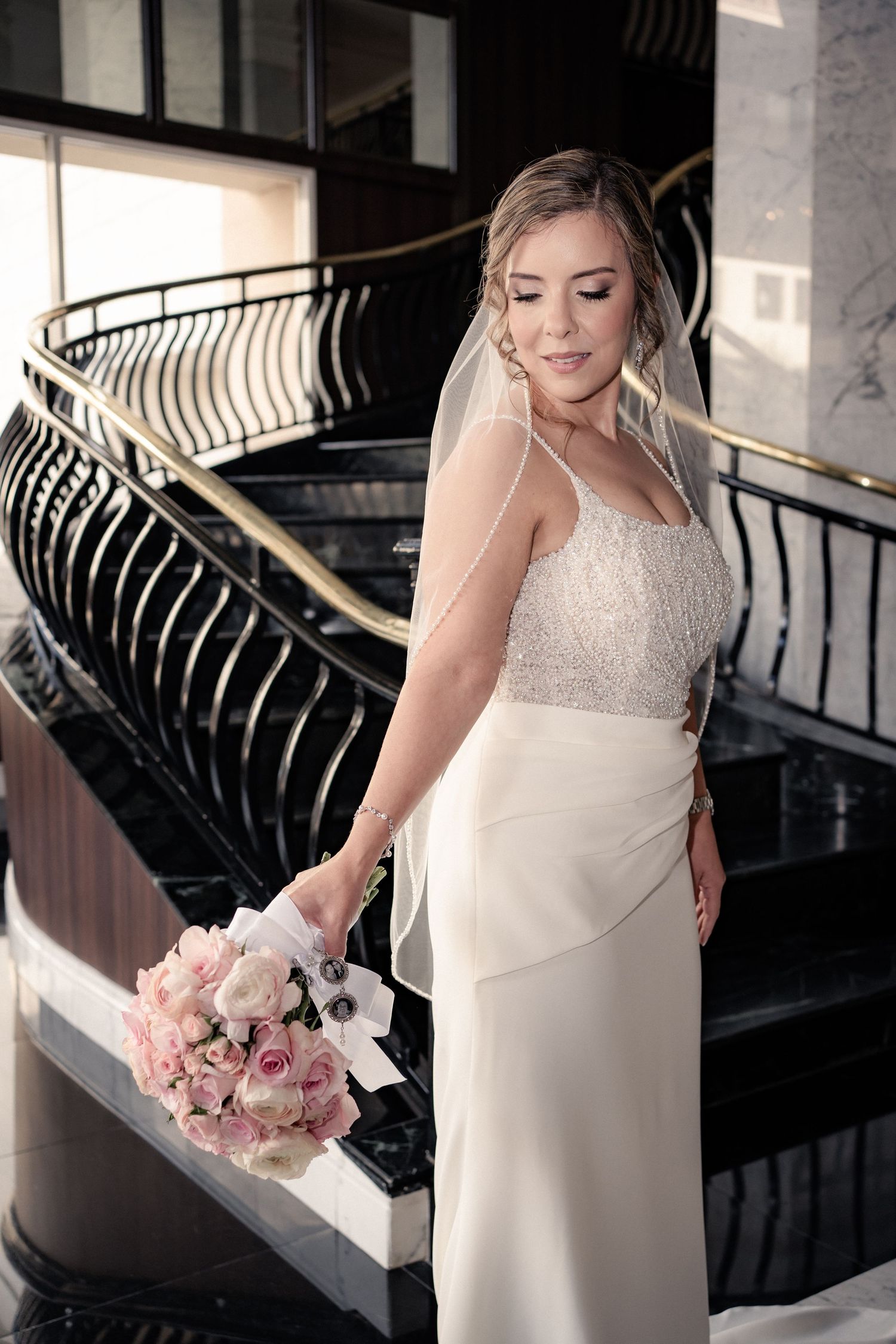 Bride in white gown and veil holds pink bouquet on elegant staircase.