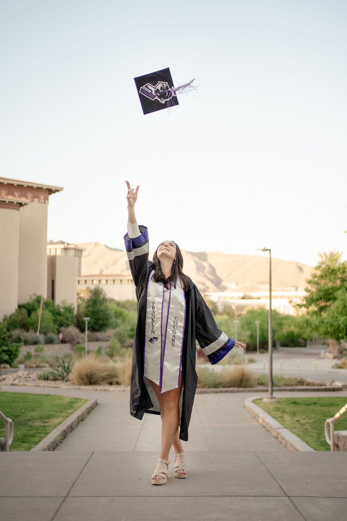 A graduate in cap and gown throws their graduation cap into the air while celebrating on a sunny campus sidewalk.