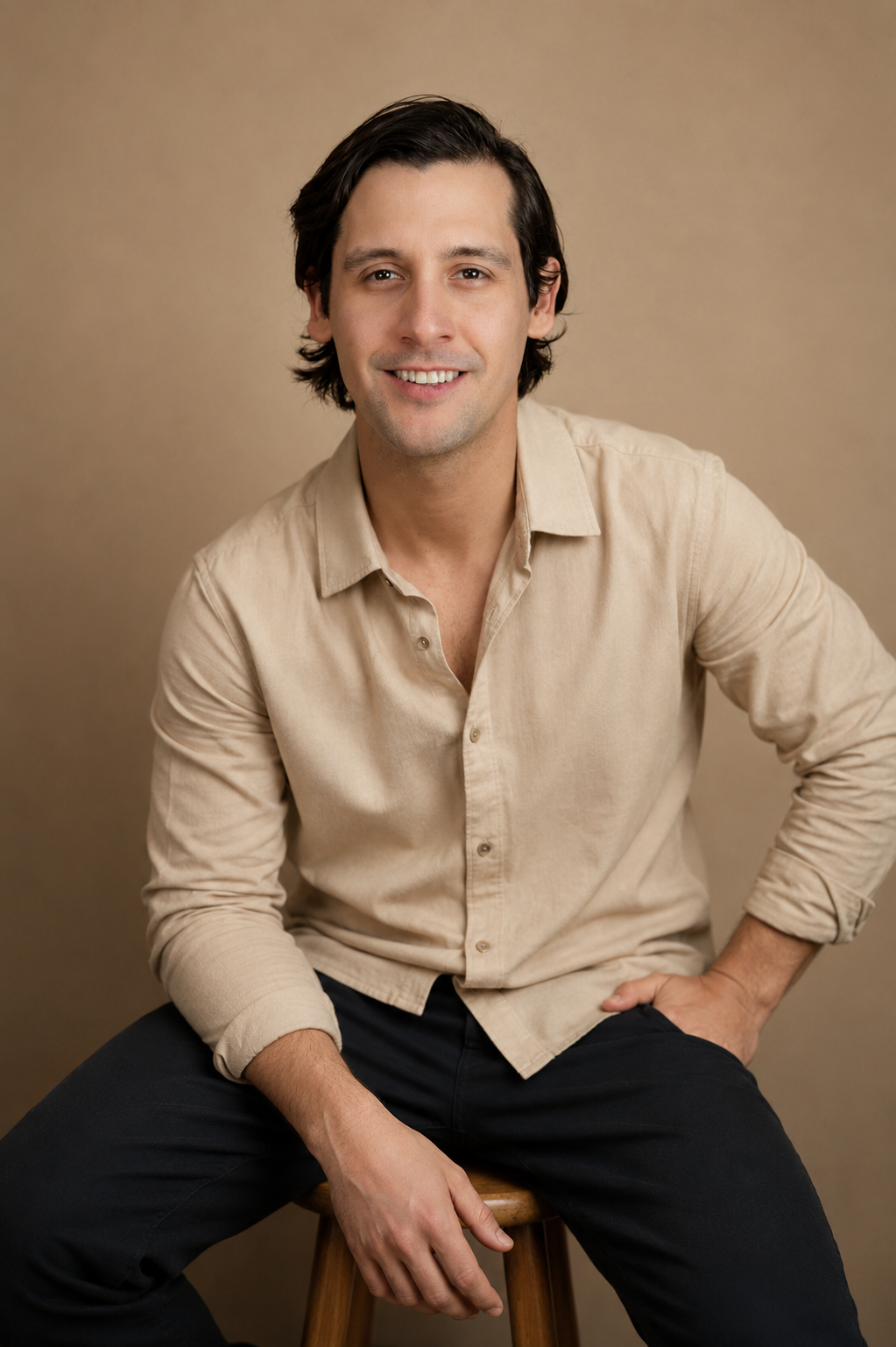 Smiling man in beige button-up shirt seated on stool against neutral tan studio background.