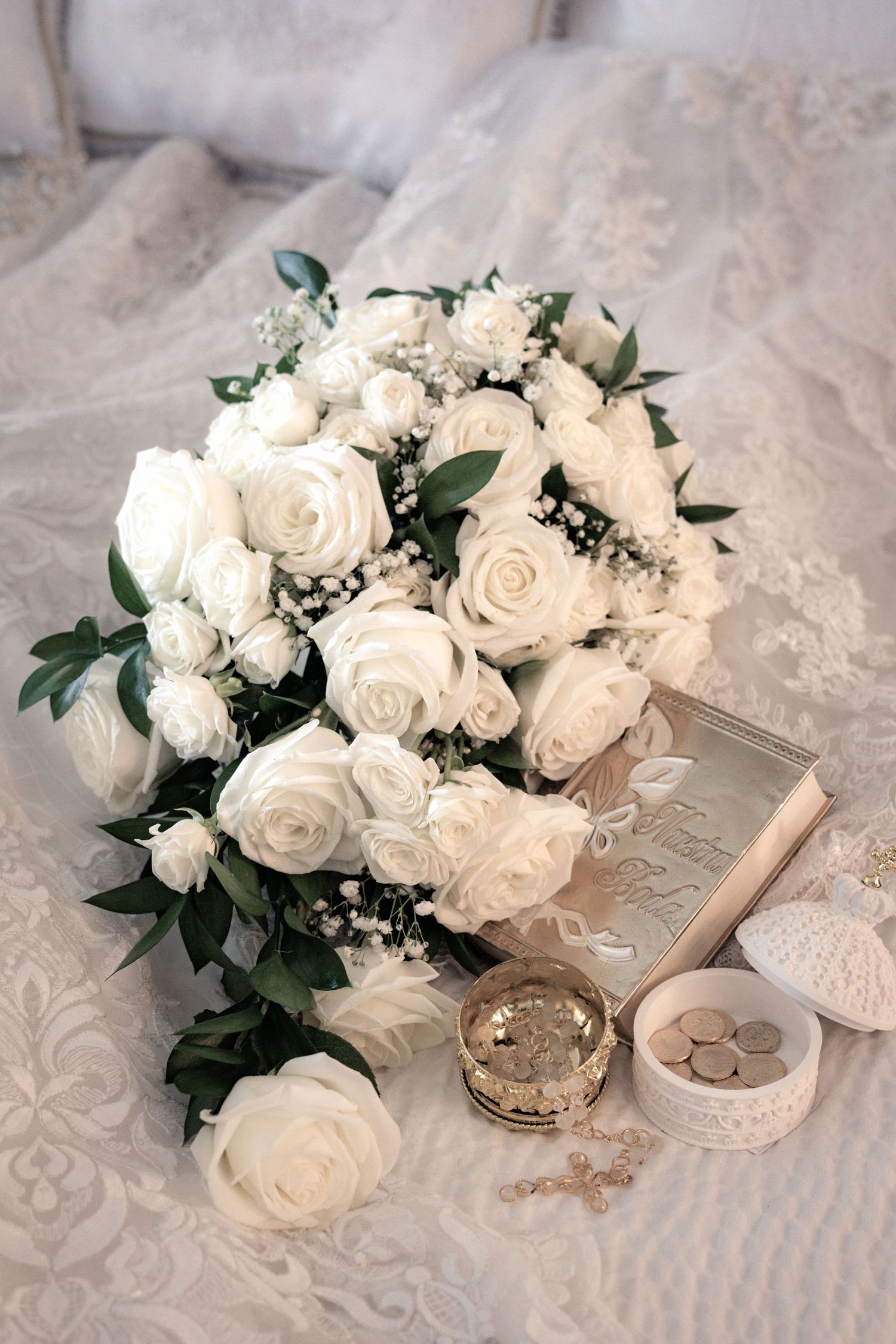 Elegant white rose bridal bouquet with greenery alongside wedding rings and delicate lace details on light fabric background.