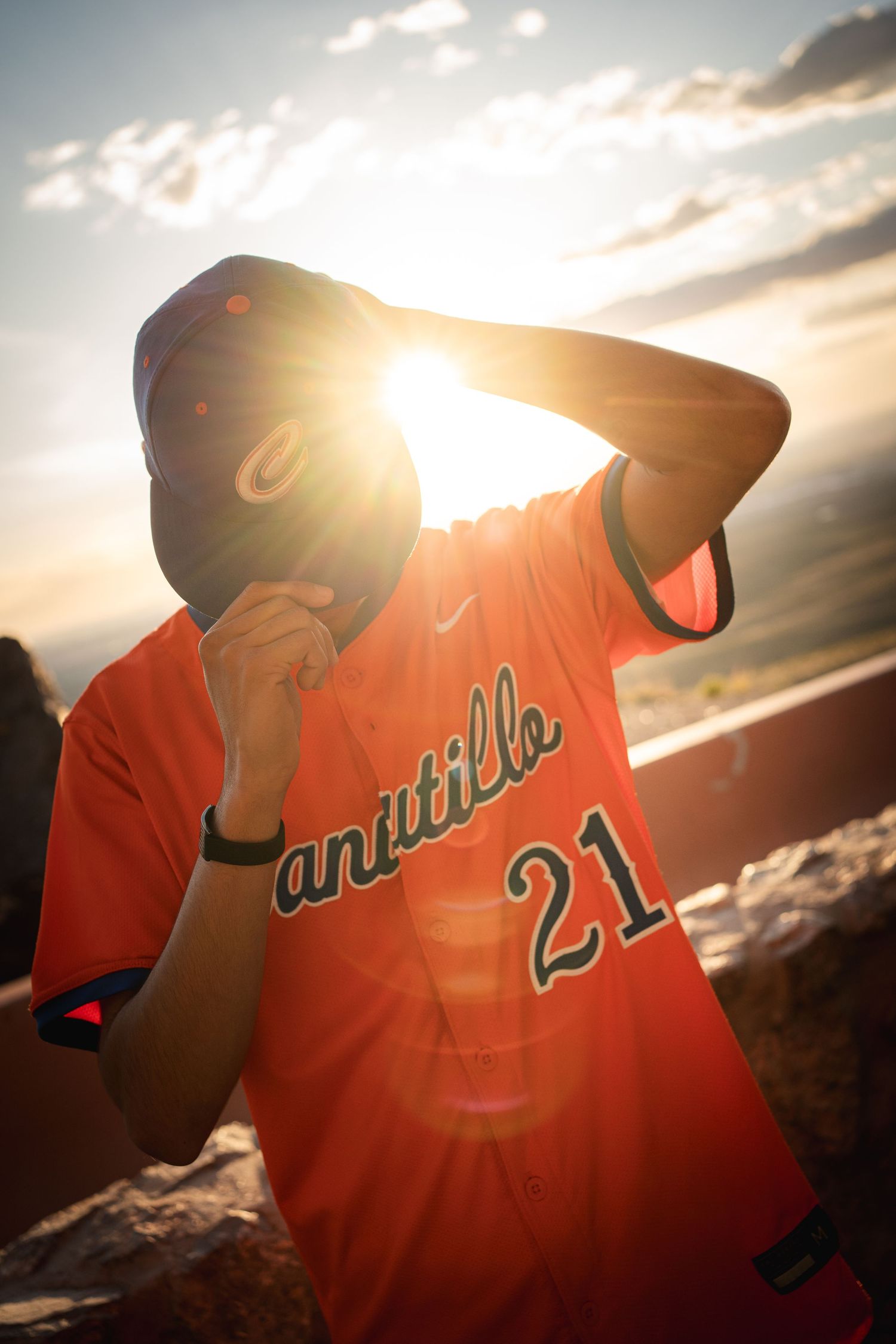 Person wearing orange Canutillo 21 baseball jersey and cap with sun flare backdrop at golden hour.