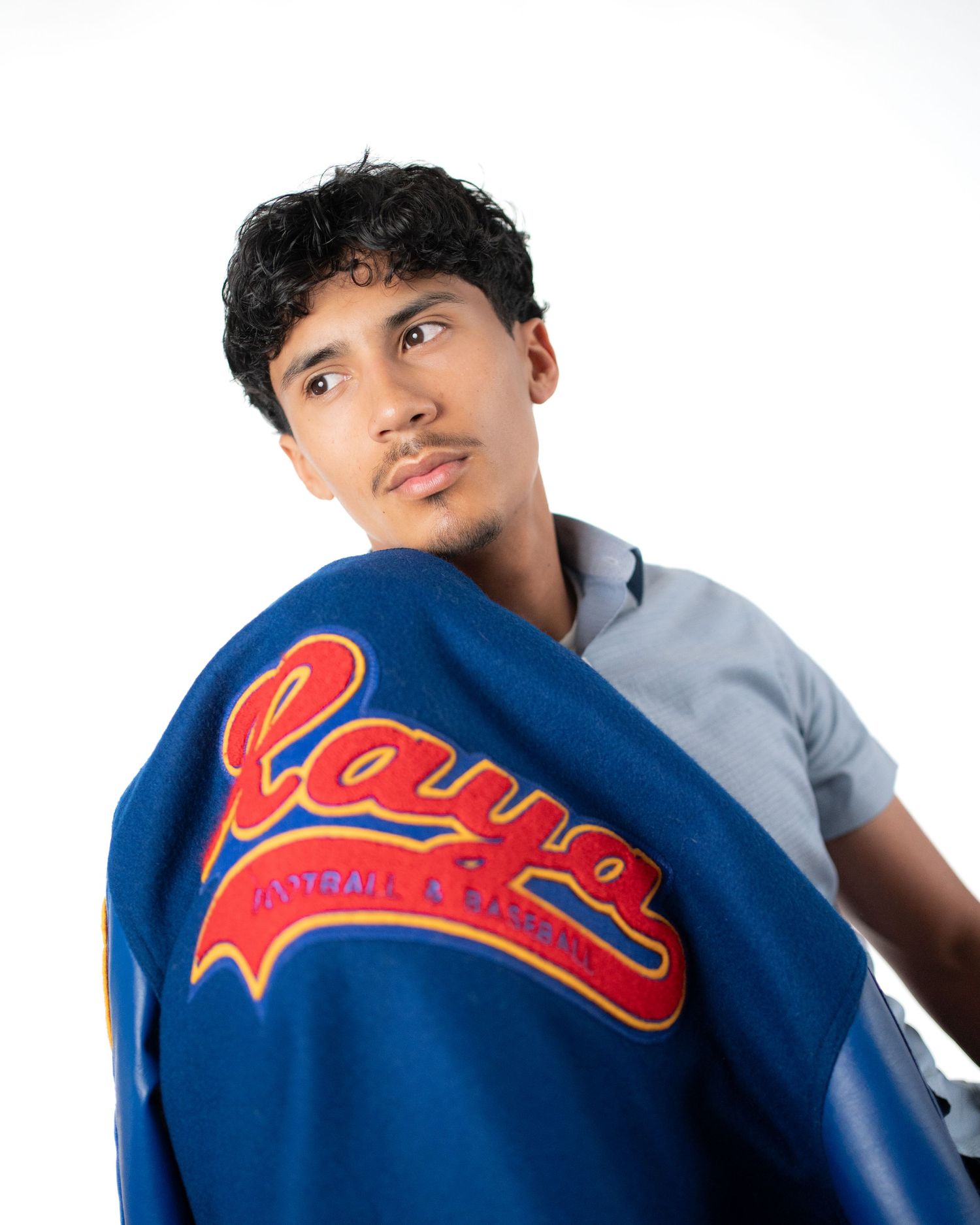 Young man in gray shirt holding blue varsity jacket with red Rayo lettering, posing against white background.