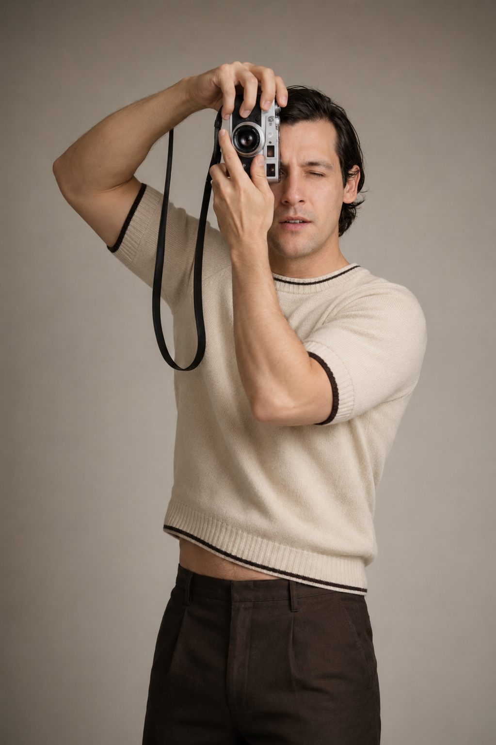 A young man in a cream sweater and brown trousers holds a vintage film camera up to his eye in a studio.