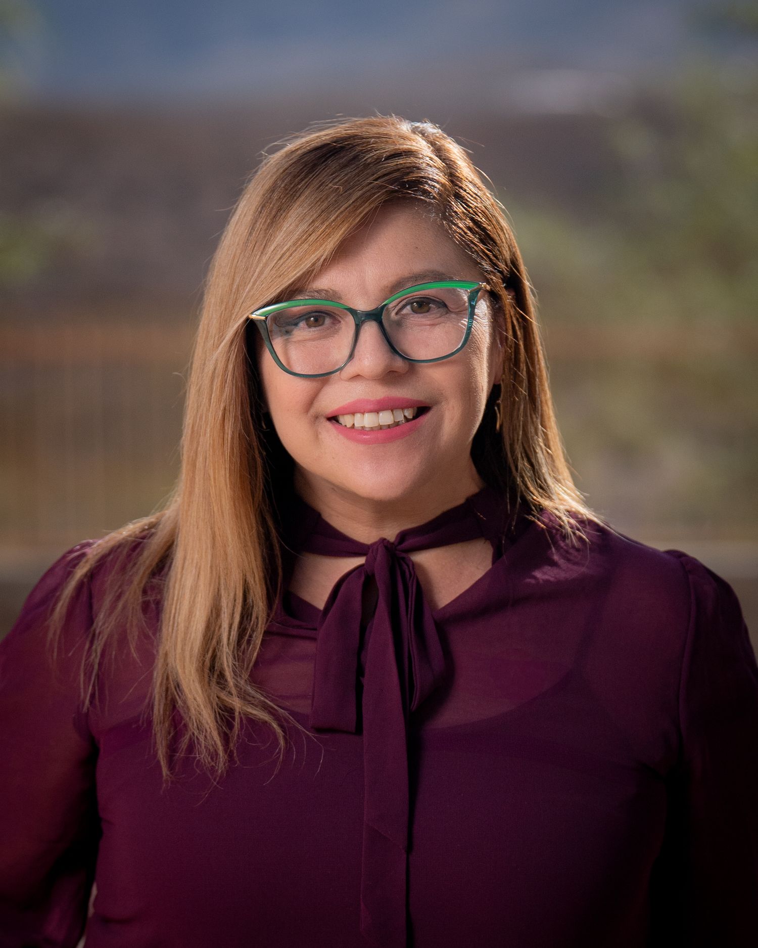 Professional headshot portrait of someone wearing green glasses and a burgundy blouse with a bow tie collar against a blurred background.