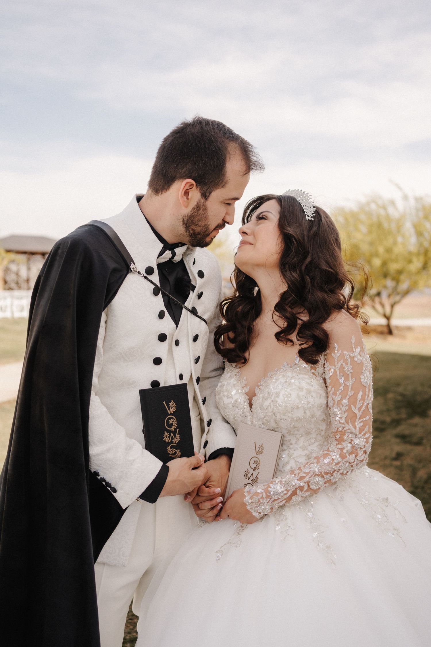 A bride and groom in elegant wedding attire share a romantic moment outdoors, holding vows books and gazing into each other's eyes.