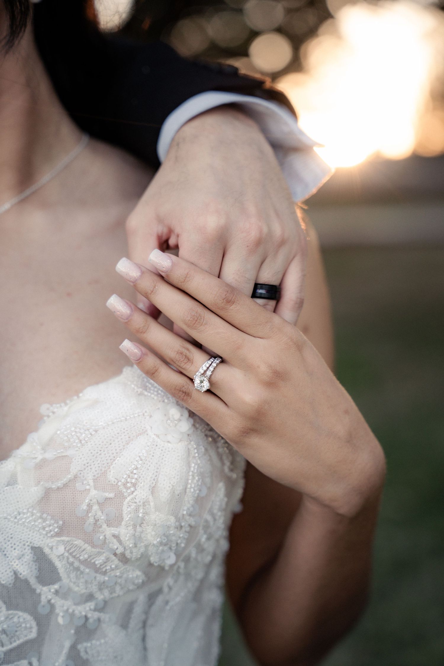 Bride and groom displaying wedding rings, bride wearing diamond band in lace dress, groom with black silicone ring at sunset.