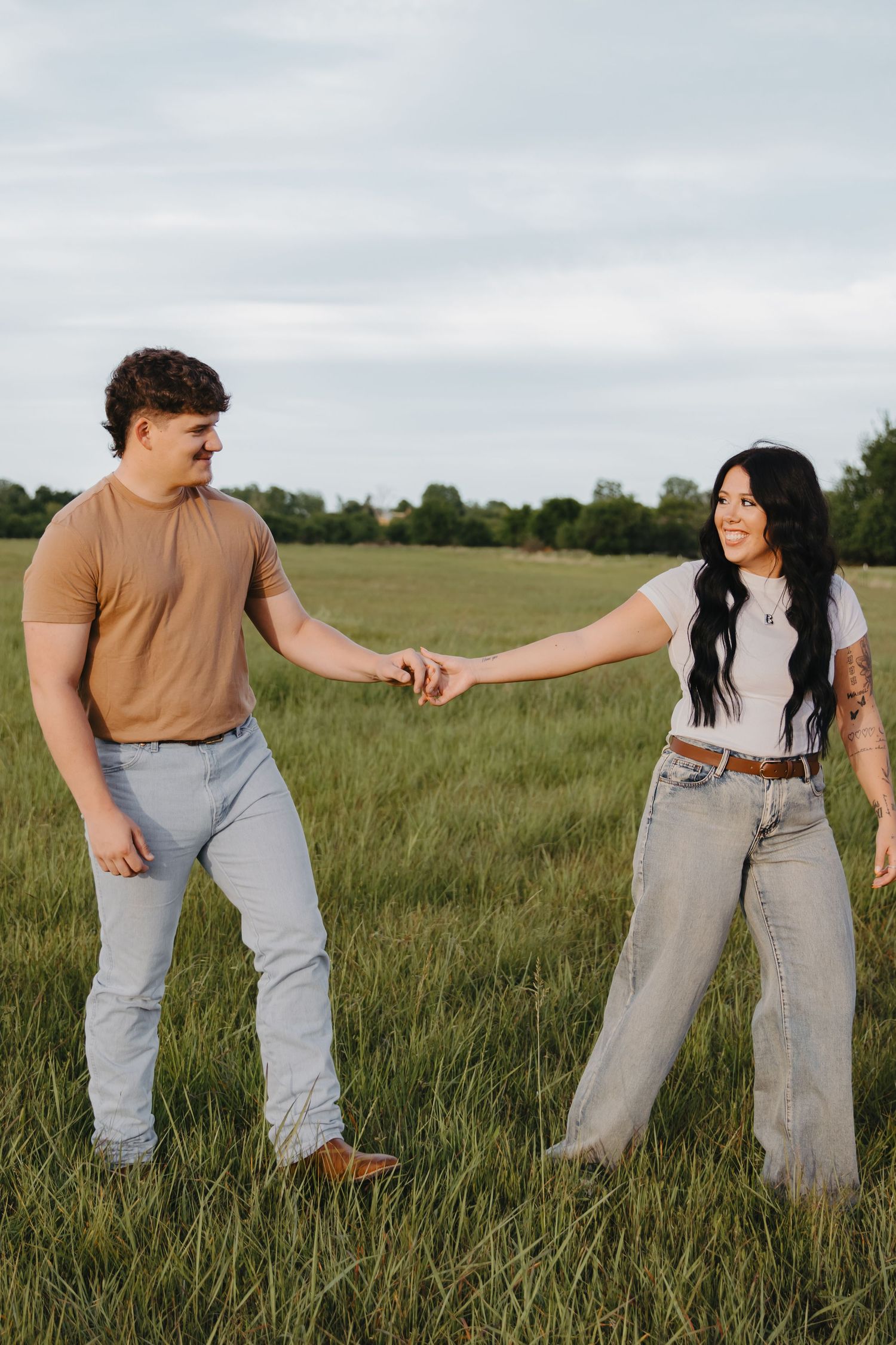 A playful couple holding hands while standing in a meadow wearing coordinated casual attire.