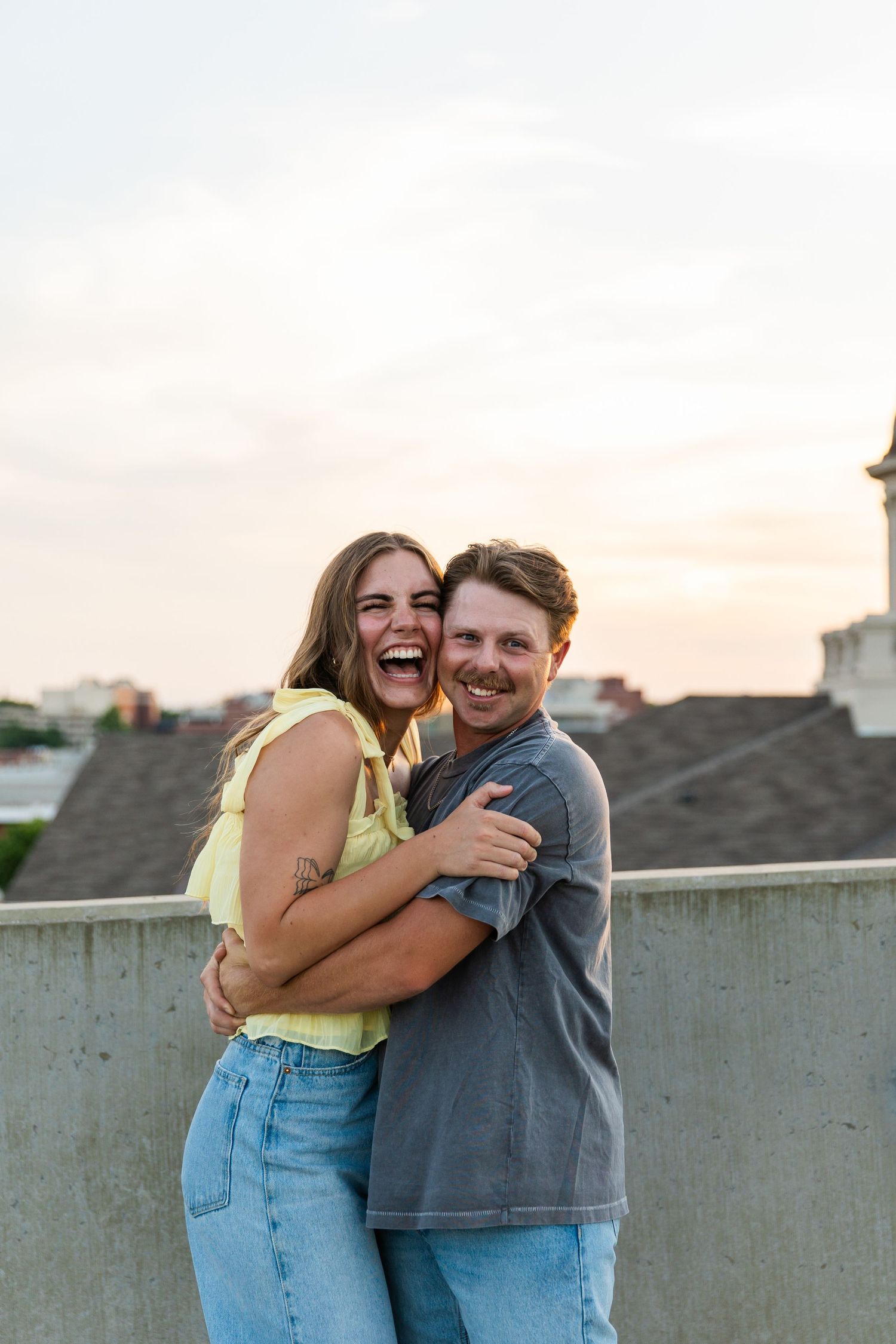 Happy couple embracing and laughing on a rooftop at sunset, wearing casual summer outfits.