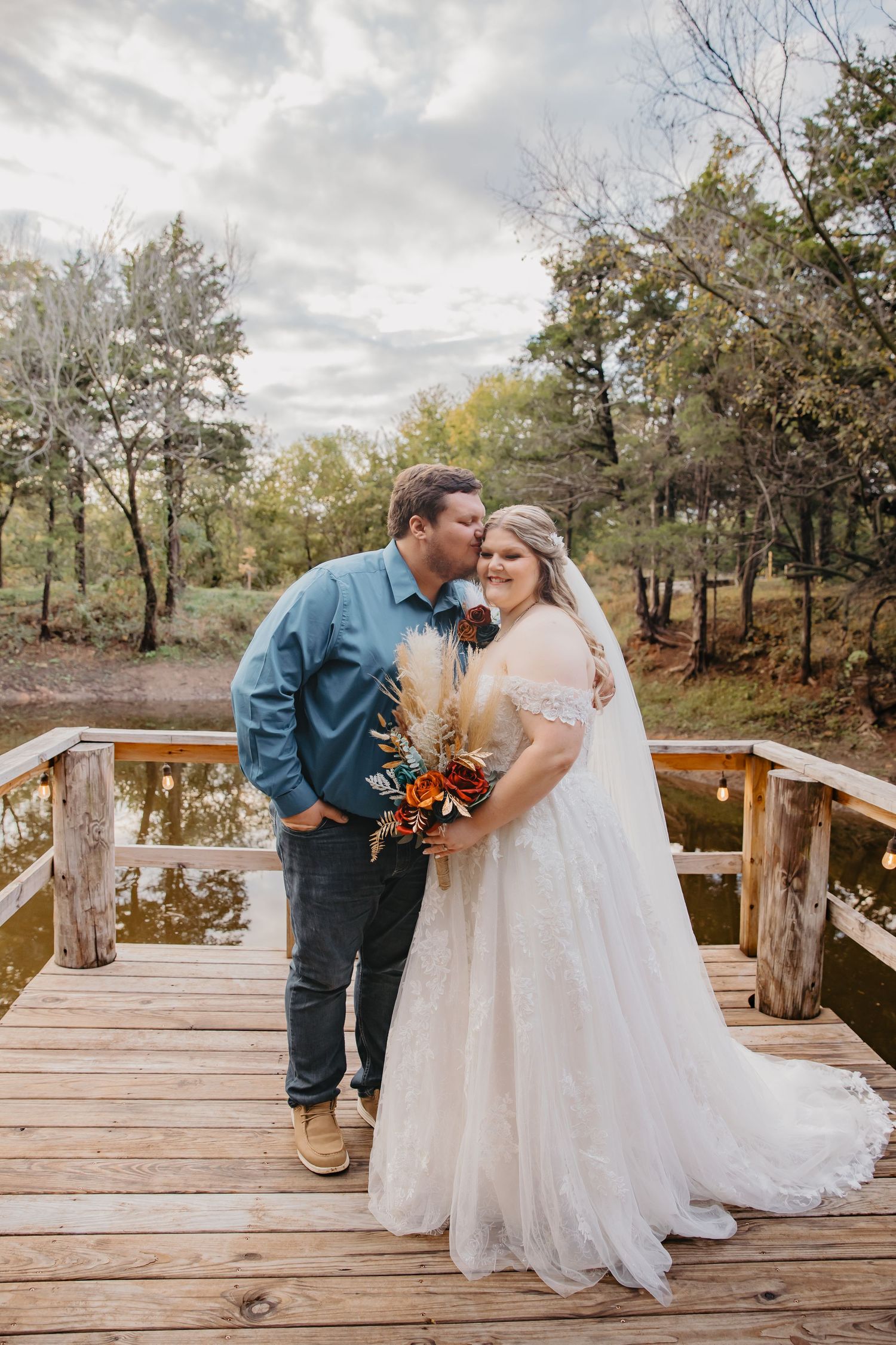 Bride and groom share a tender kiss on a wooden bridge surrounded by autumn trees and a serene pond.
