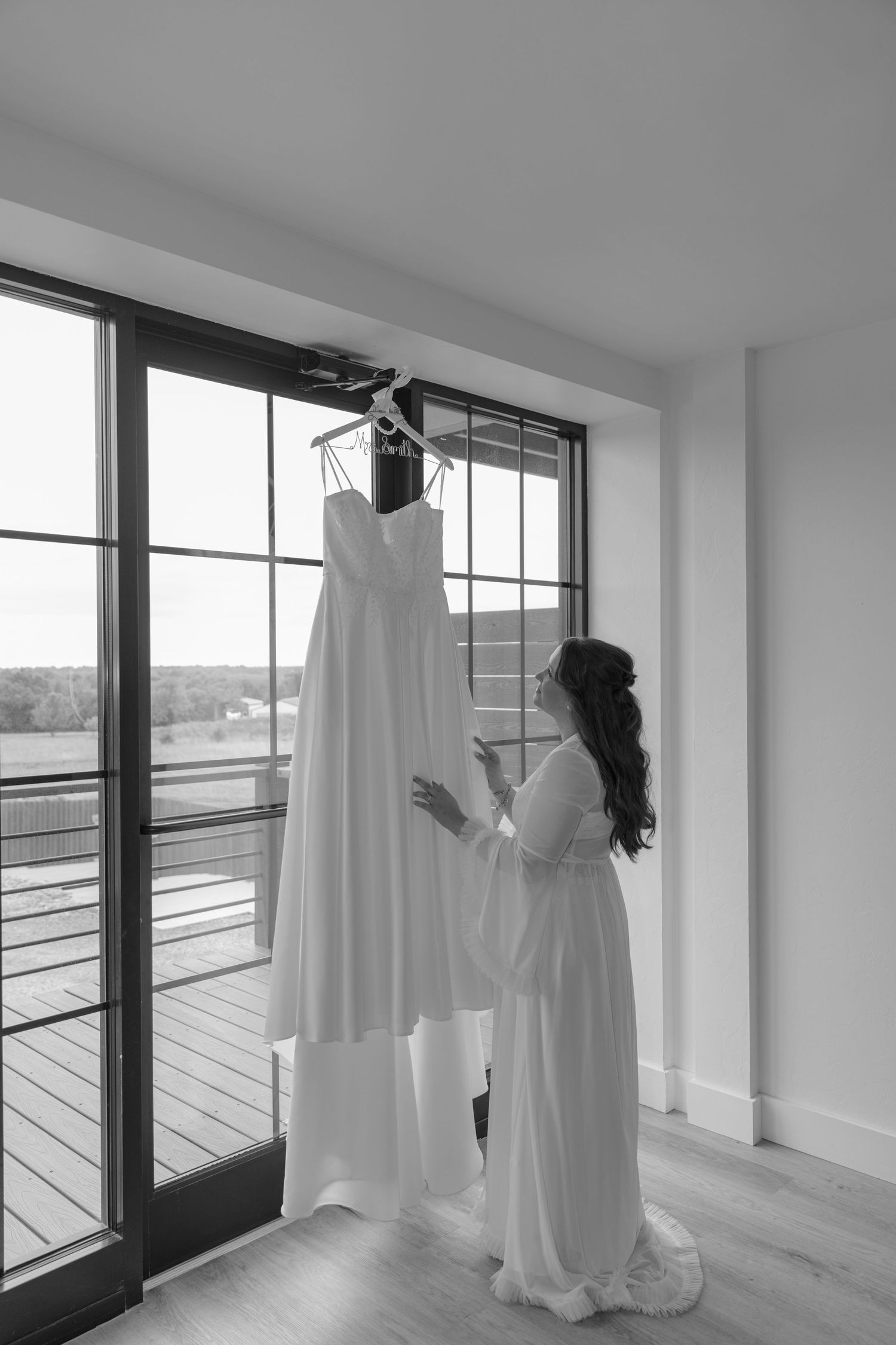 Bride in robe admiring her wedding dress hanging by a window in black and white bridal preparation photo.