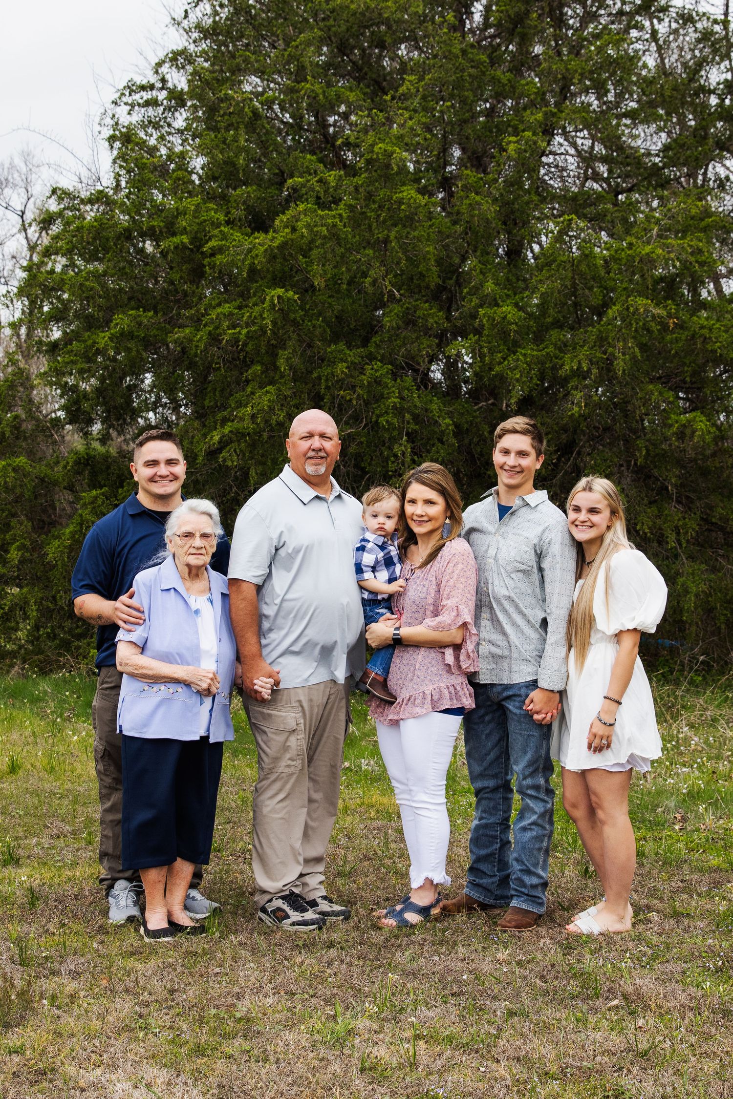 Multi-generational family portrait outdoors with large tree in background.
