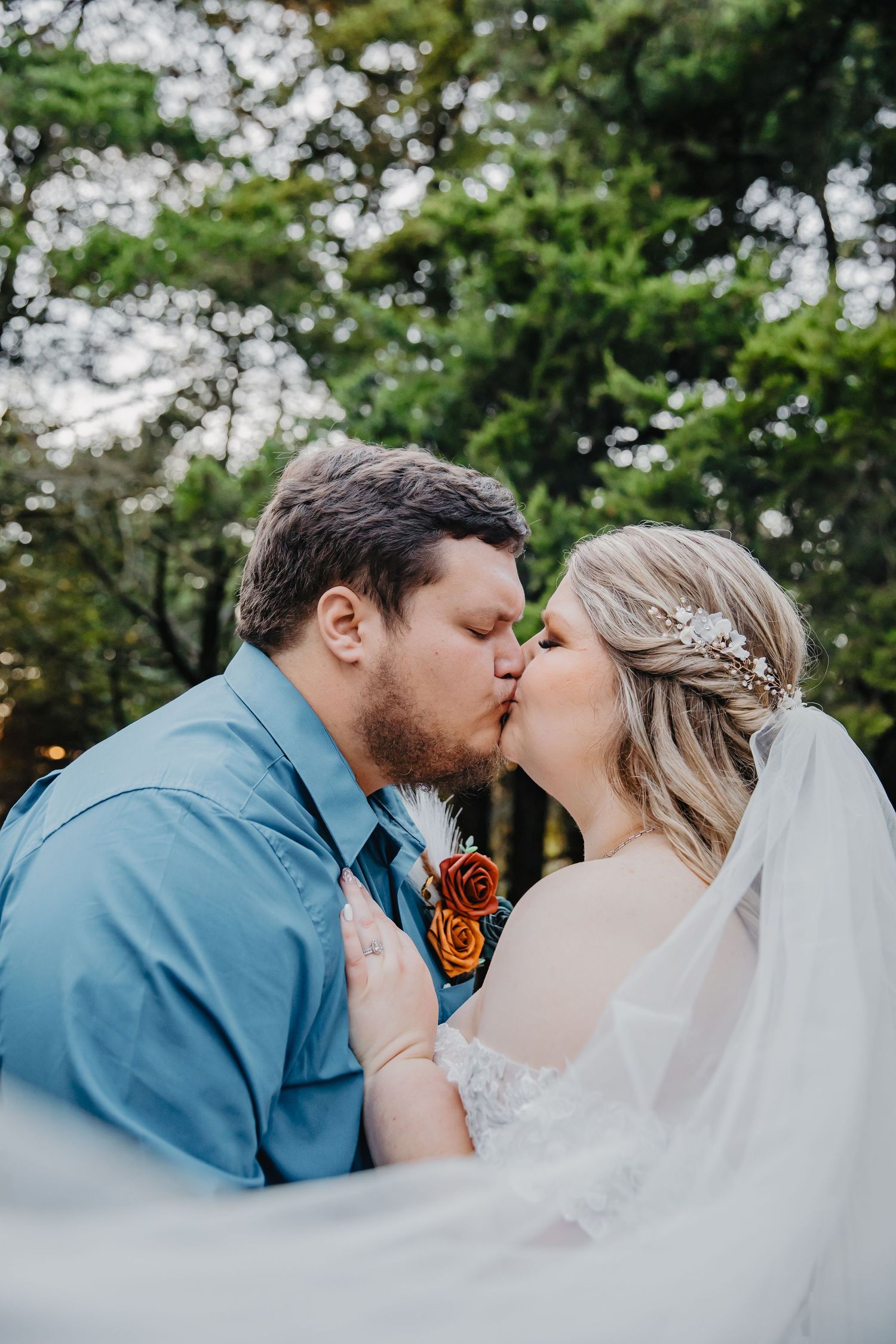 Bride and groom share a romantic kiss outdoors, bride wearing lace gown with floral crown and flowing veil.