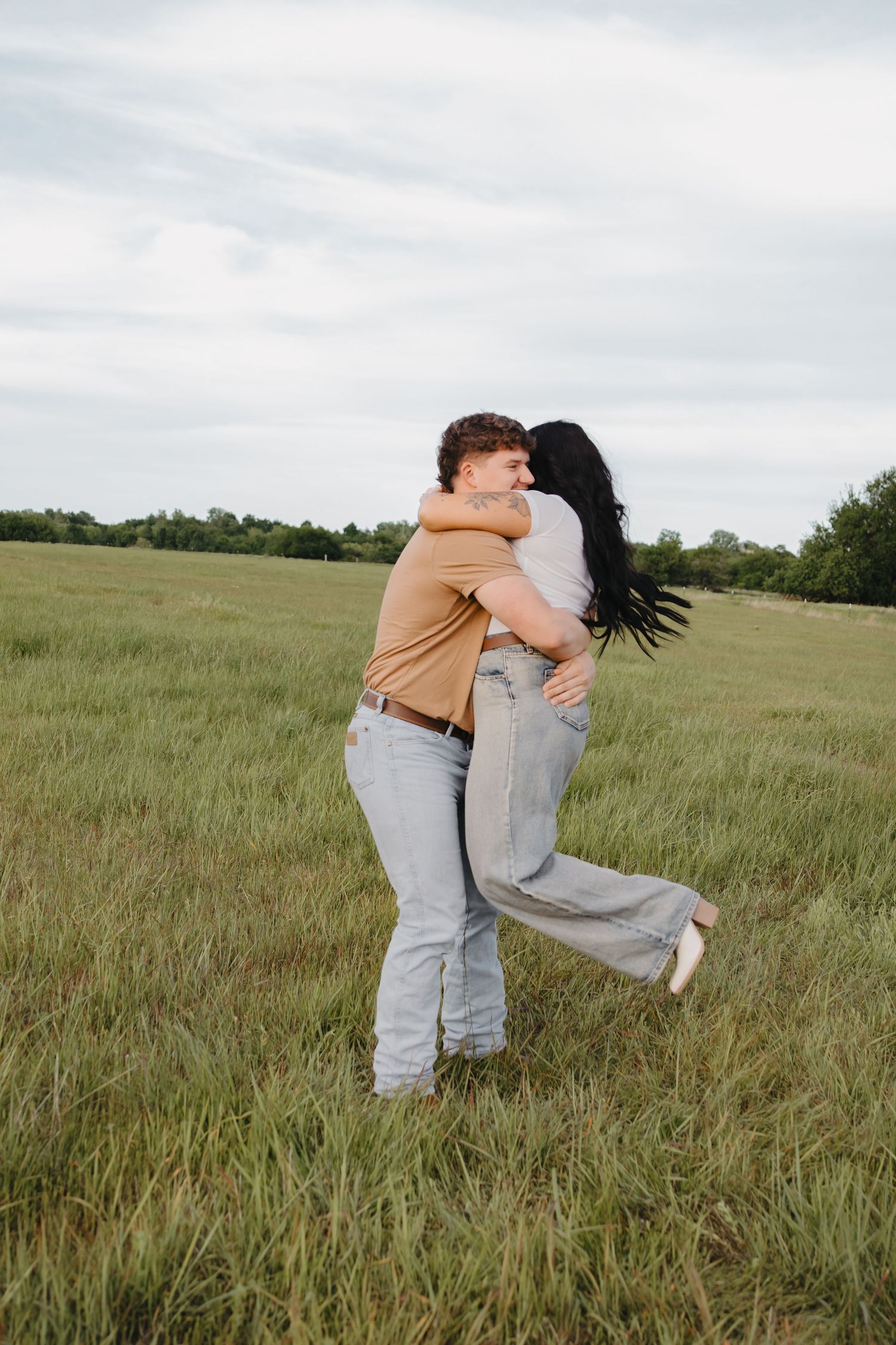 A couple embracing in an open field with tall grass and cloudy skies.