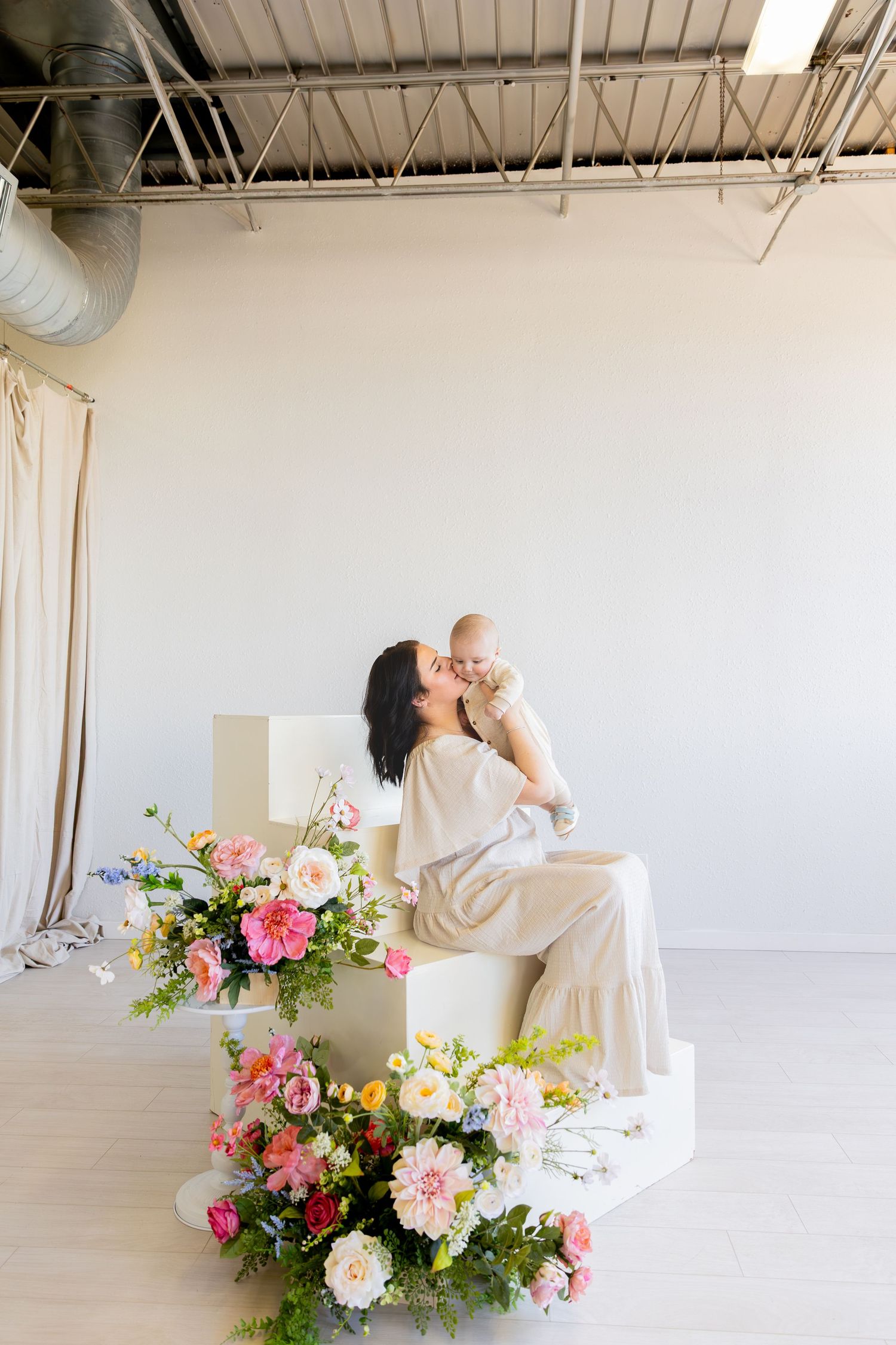 A person in white loungewear sits on steps surrounded by vibrant pink and white floral arrangements in a bright studio space.