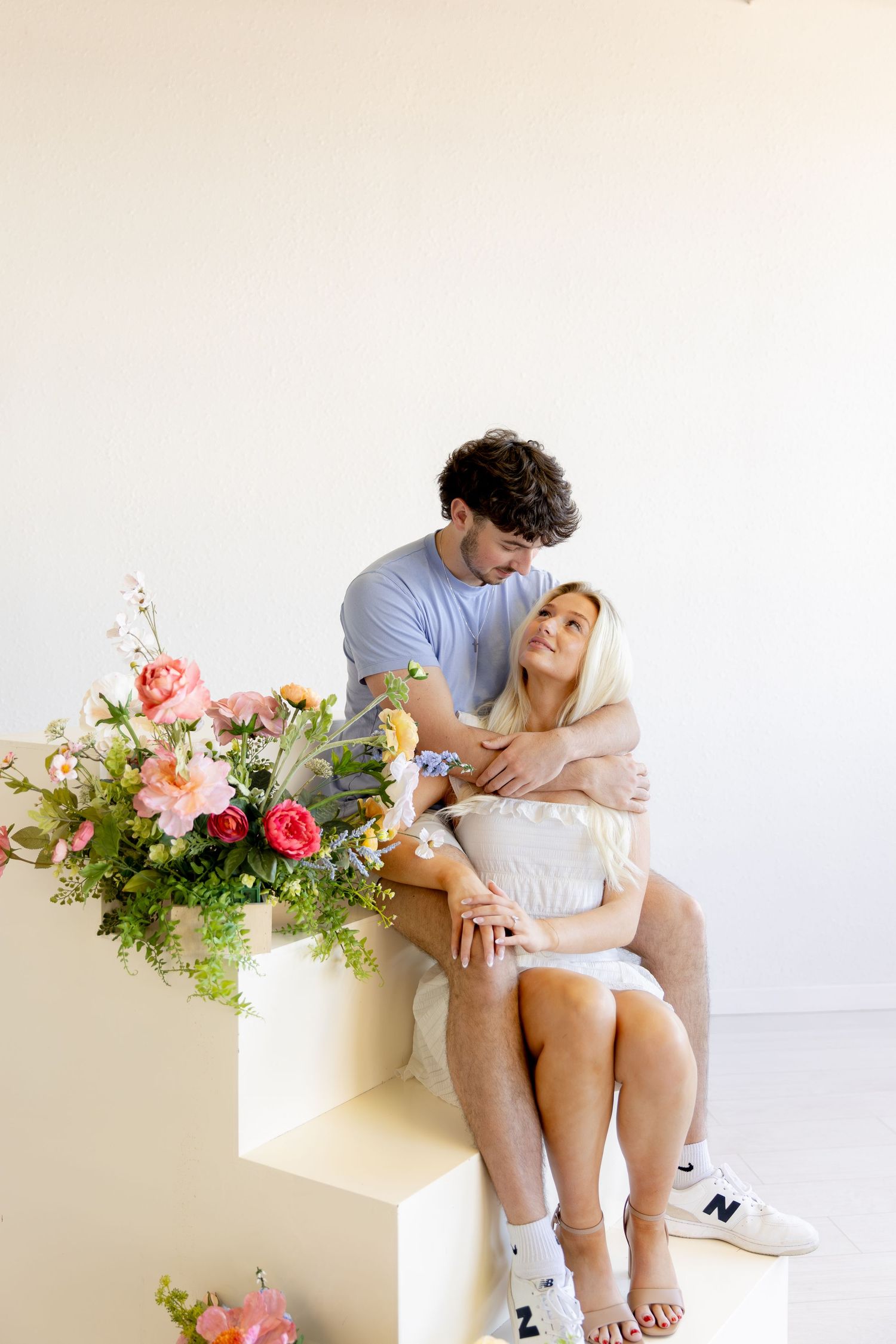 A couple sits together on a white bench with pink floral arrangements in a minimalist setting.