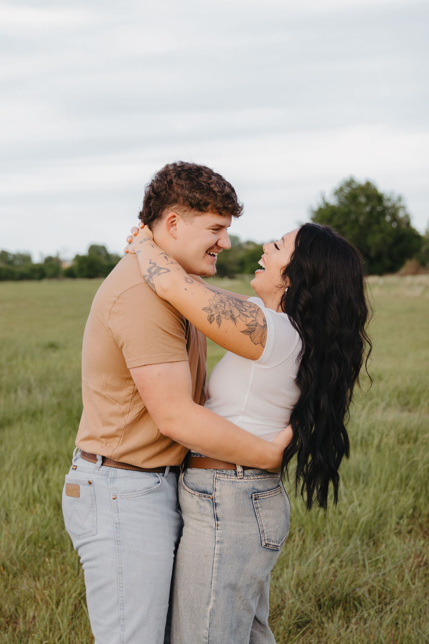 A couple embraces and laughs together in a sunlit meadow wearing casual attire.