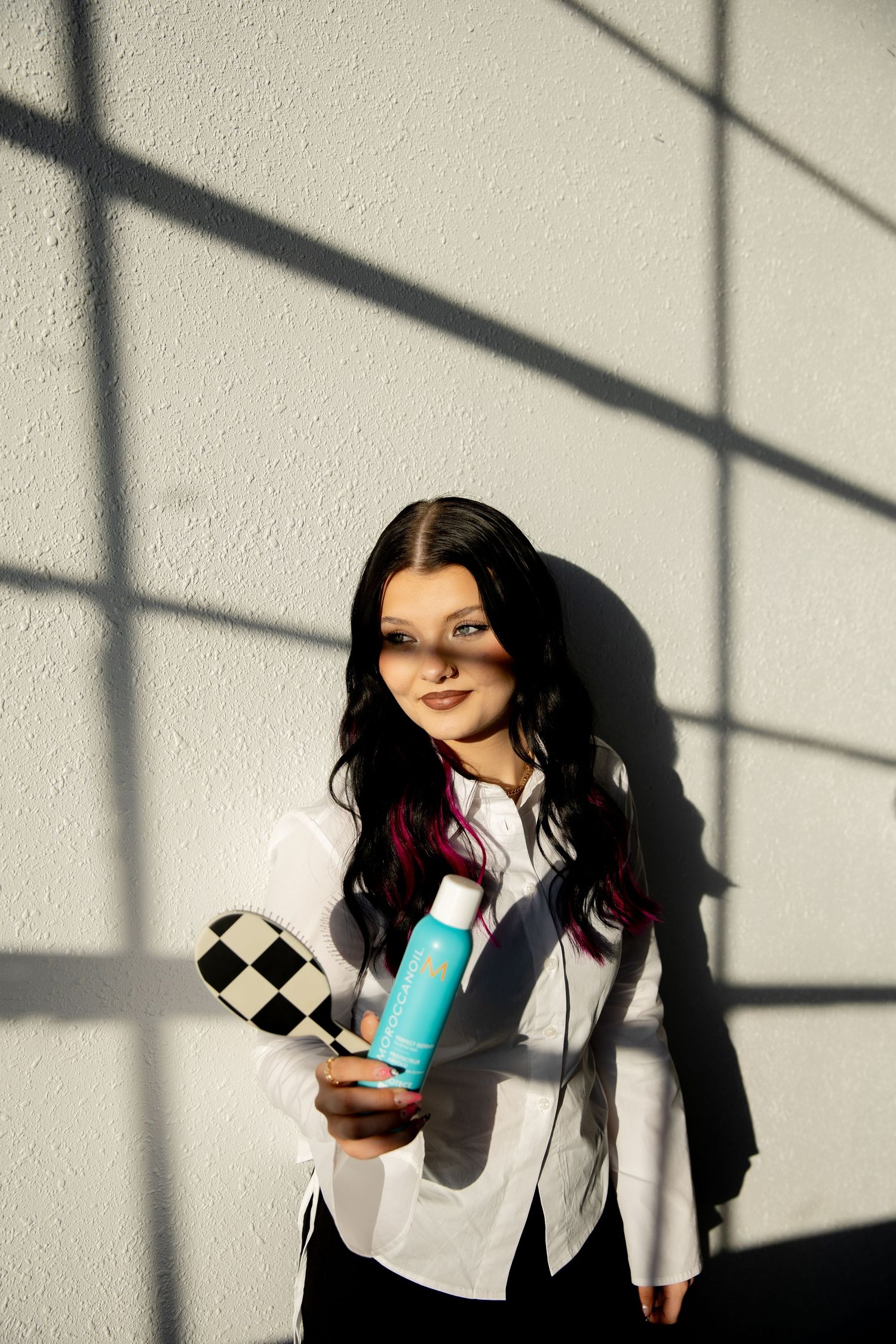 Young woman with dark hair holding a turquoise hair product bottle and checkered paddle against a white wall.