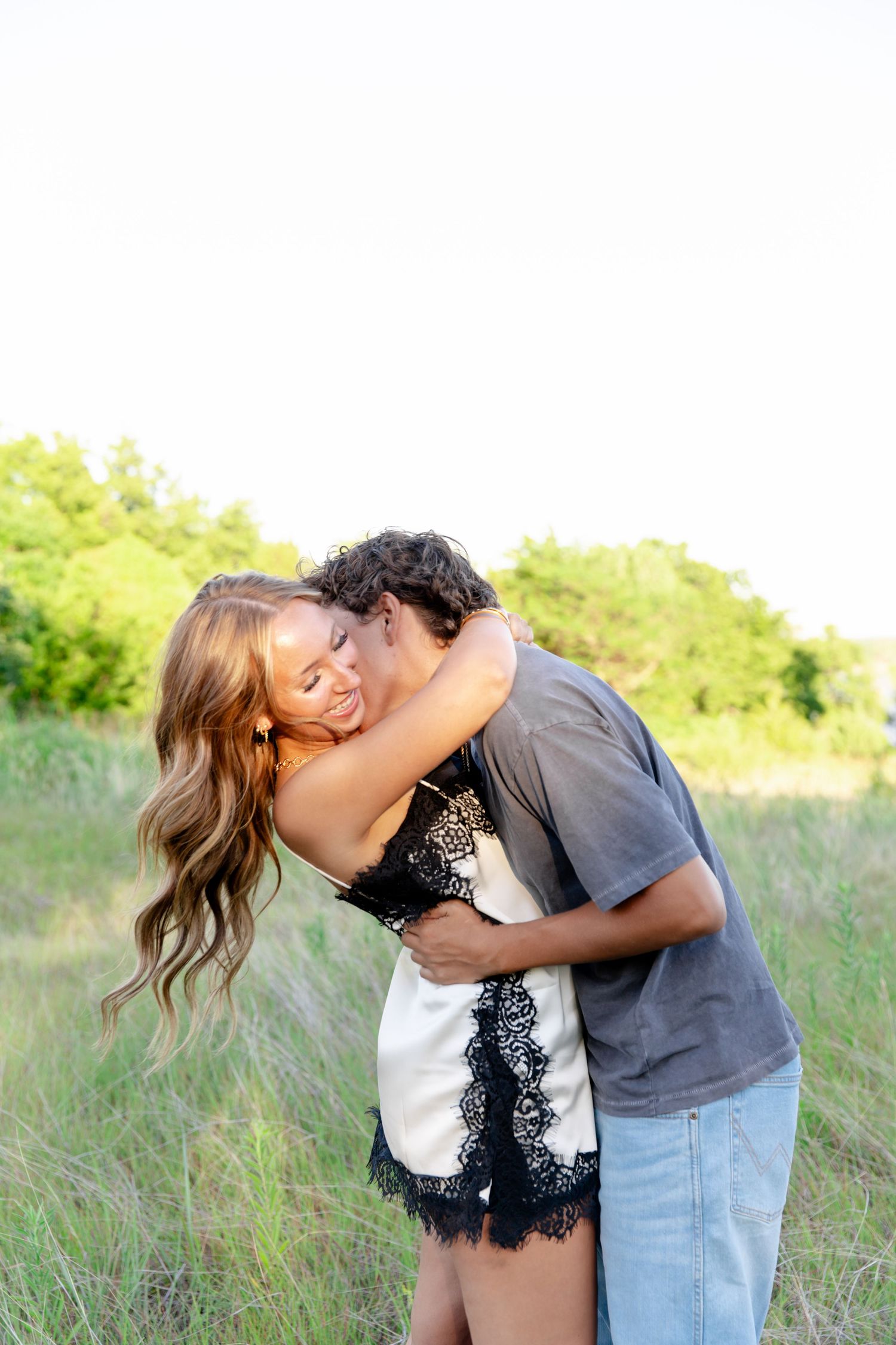 Couple embracing romantically in a green meadow, woman in black lace dress smiling as man kisses her cheek.