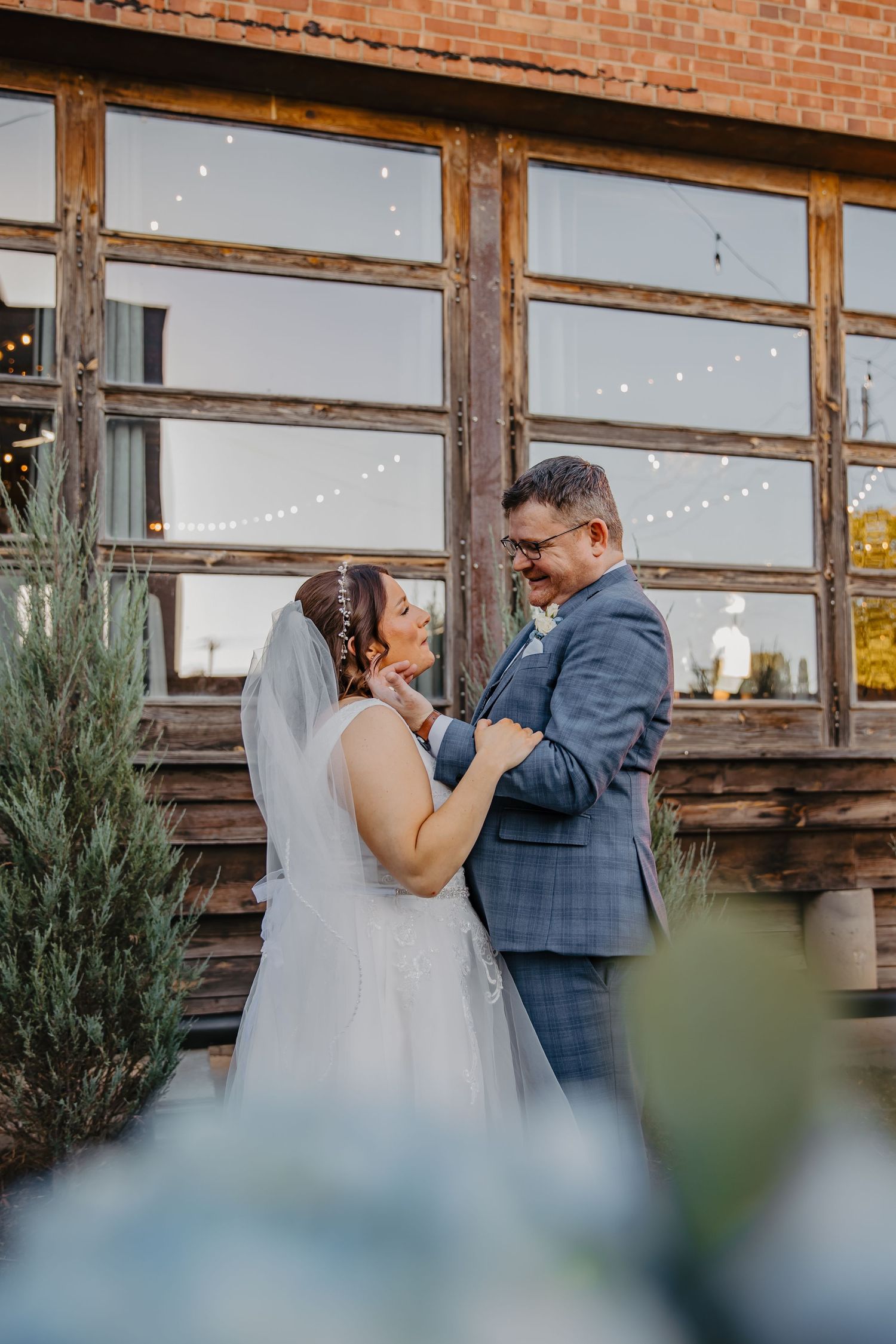 A bride and groom share a joyful moment outside a rustic venue with string lights, dressed in a wedding gown and blue suit.
