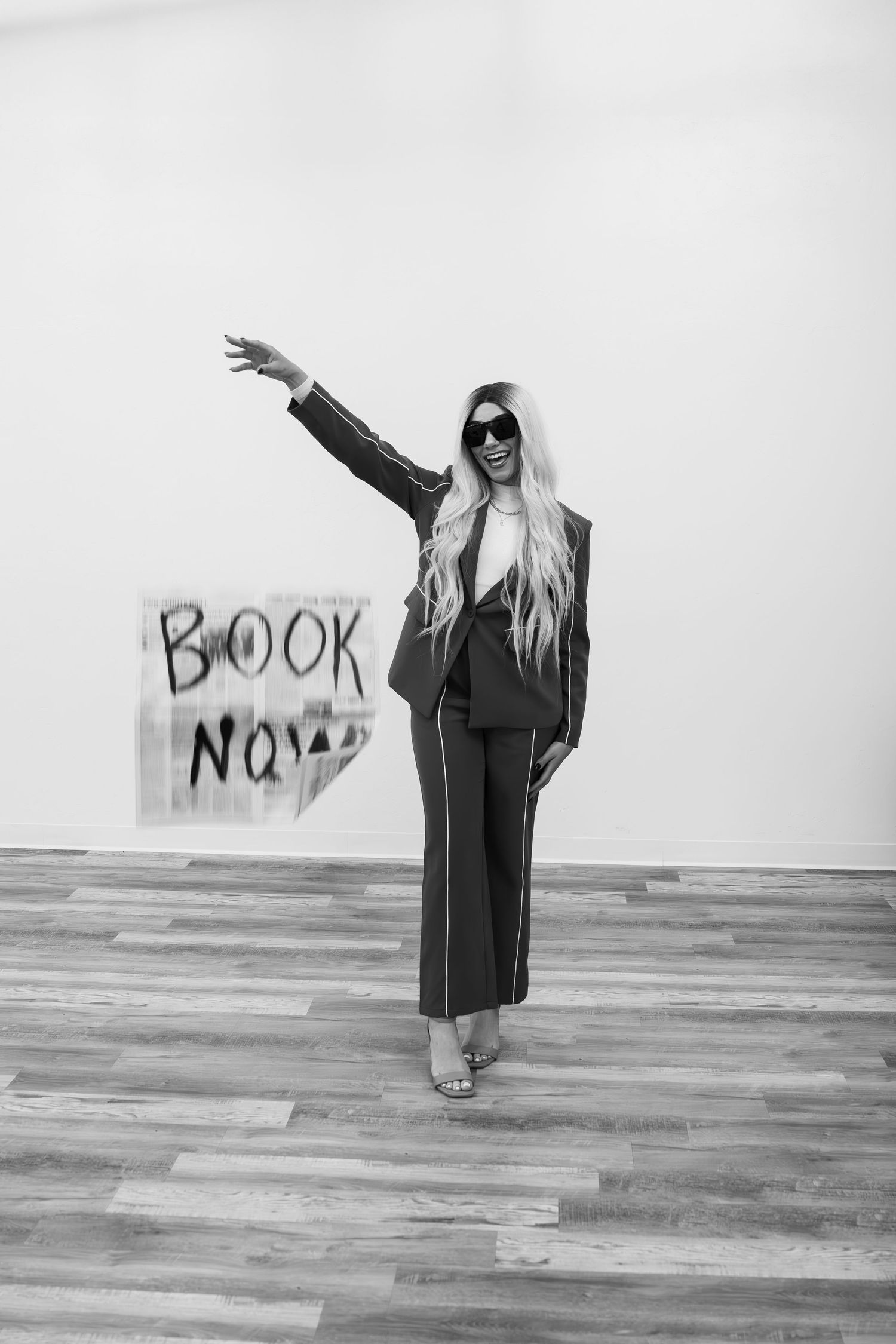 Person in black business attire poses with raised arm next to 'Book Now' text against white wall.