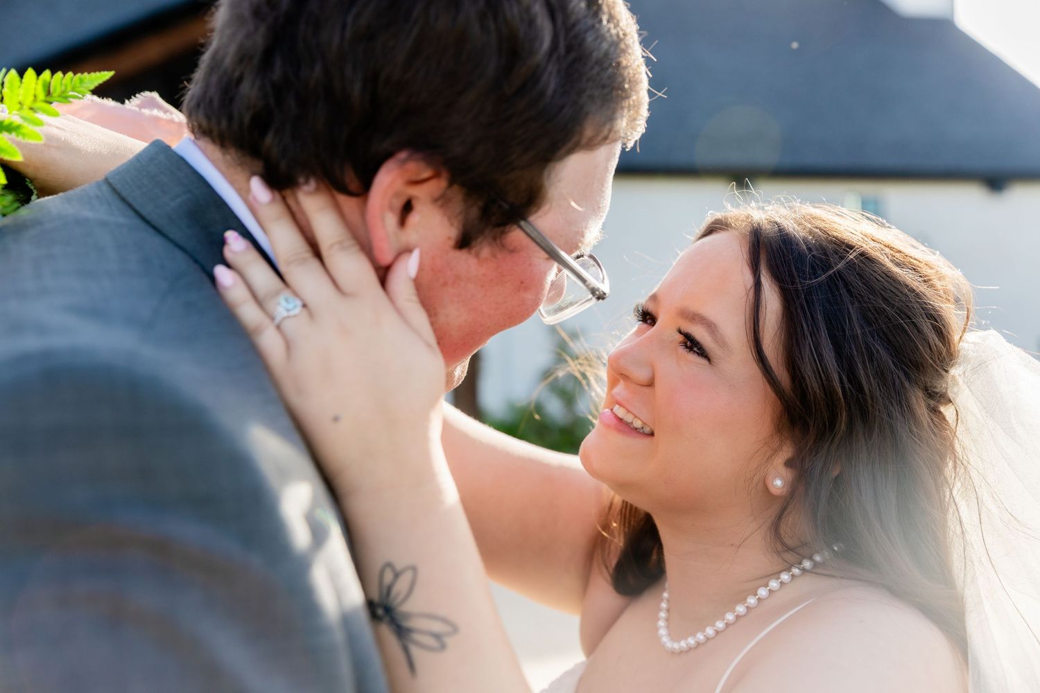 Bride smiling up at groom during outdoor wedding ceremony, bride wearing pearl necklace and veil, groom in gray suit.