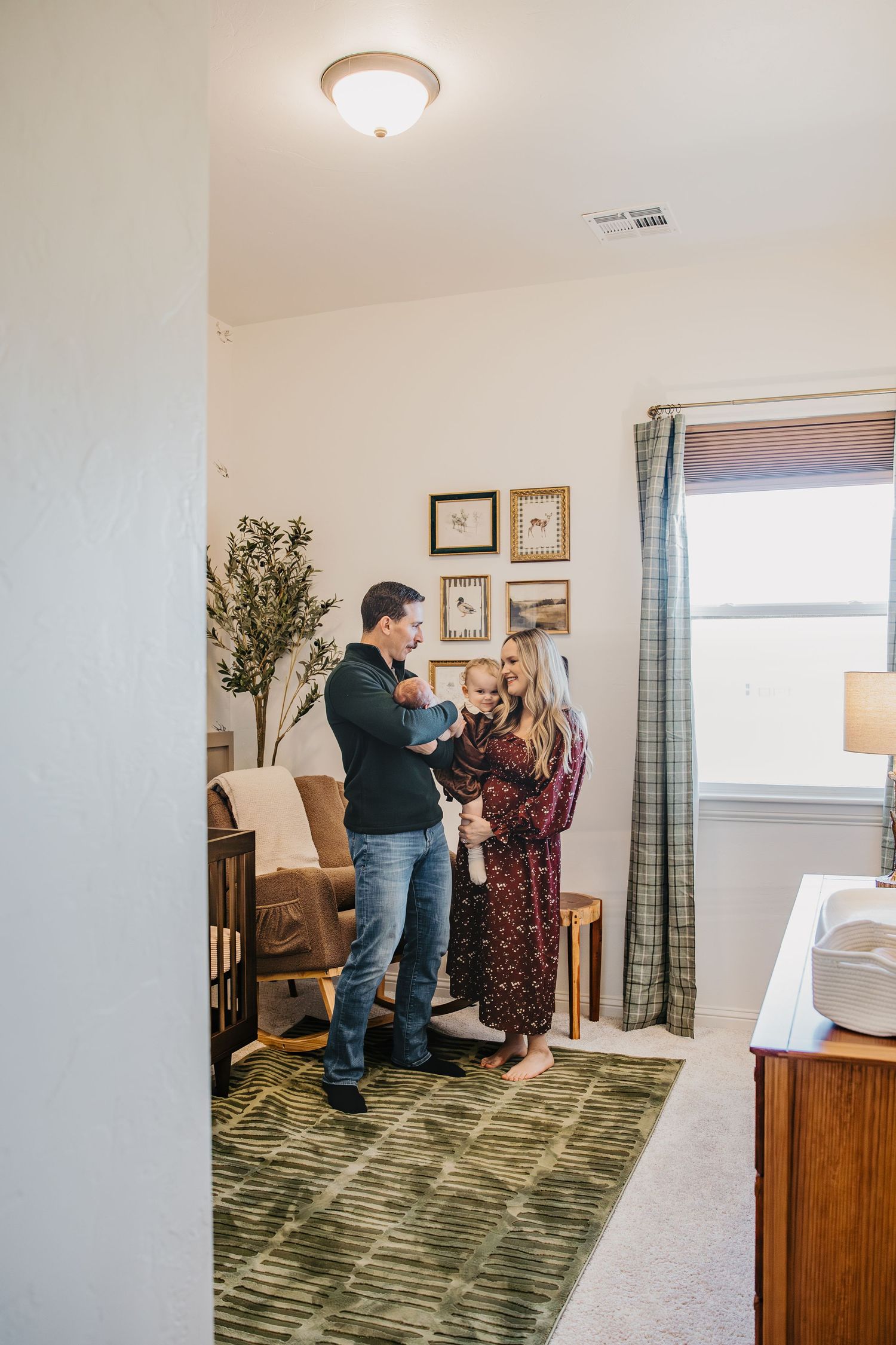 A young family stands together in a cozy nursery, parents holding their newborn baby near a crib and gallery wall.