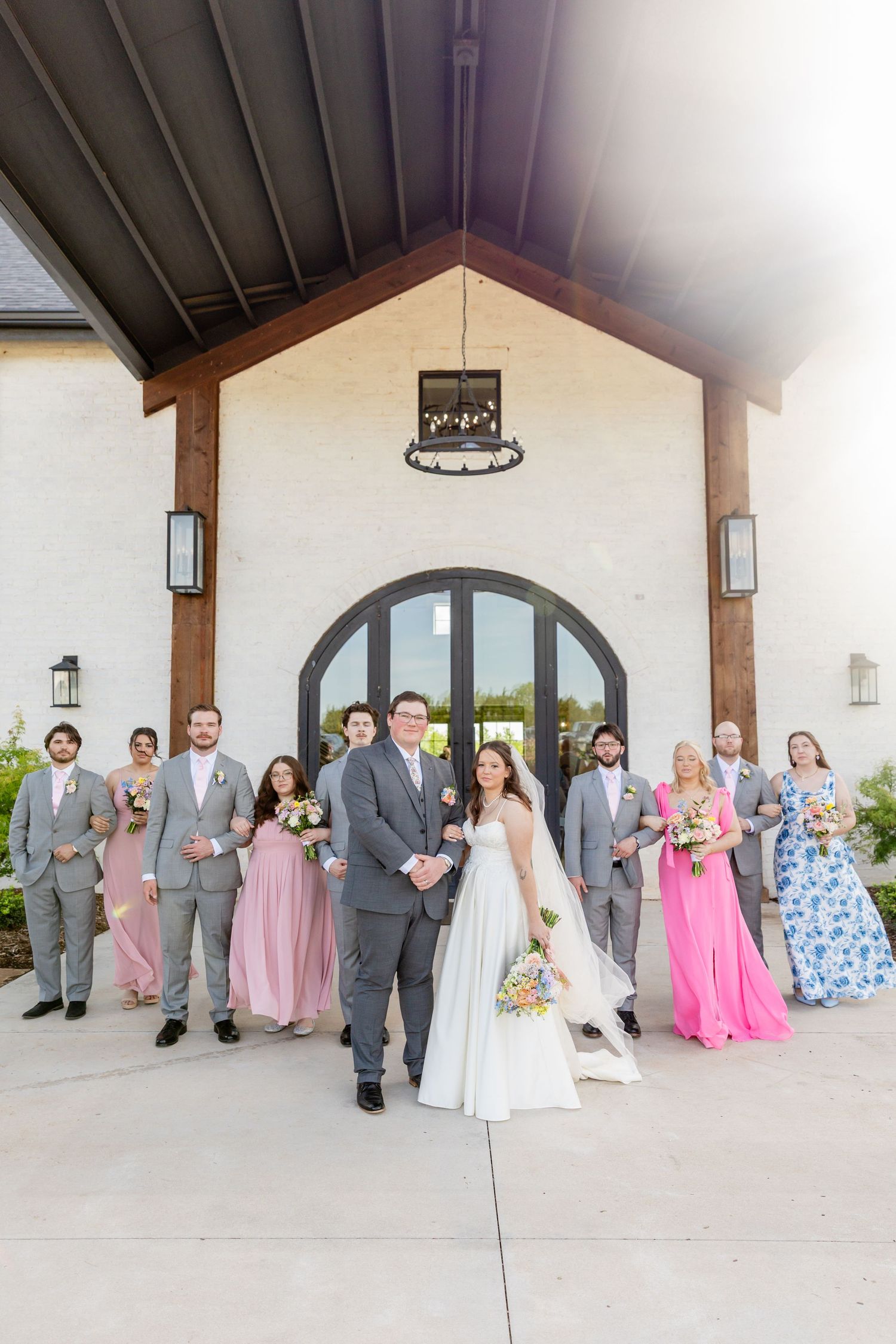 Wedding party poses outside a white brick venue, bride and groom centered with bridesmaids in pink and groomsmen in gray.