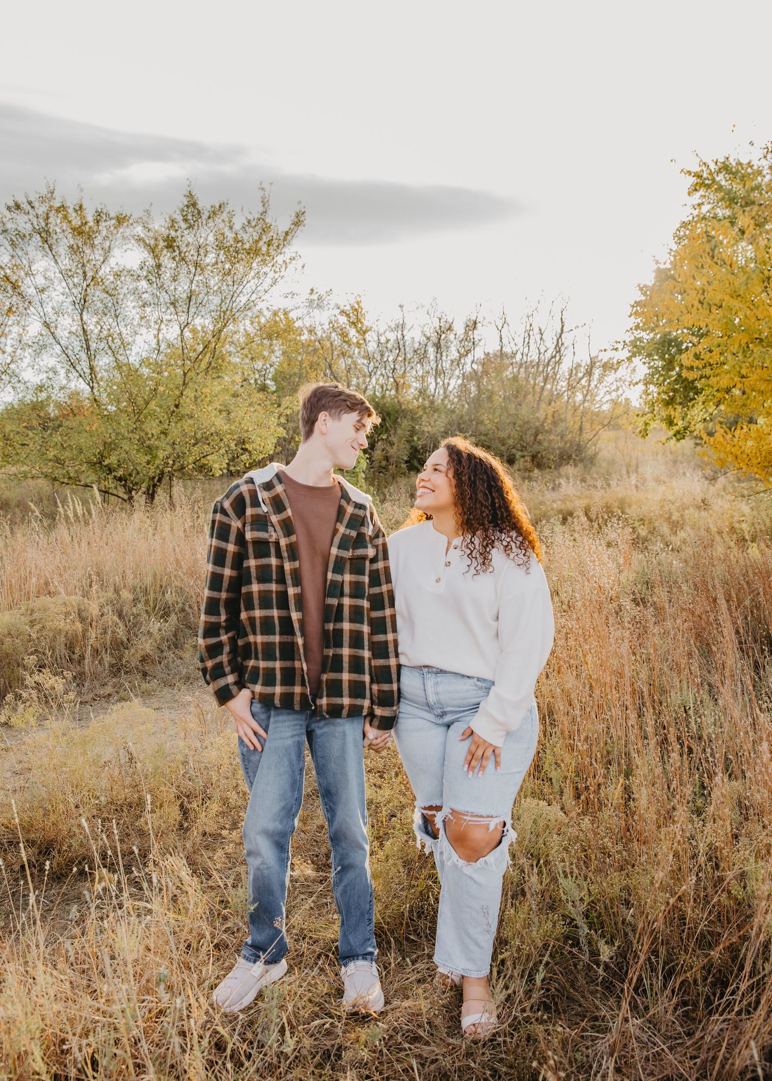 A young couple stands together in a golden autumn field, smiling at each other in casual fall outfits.