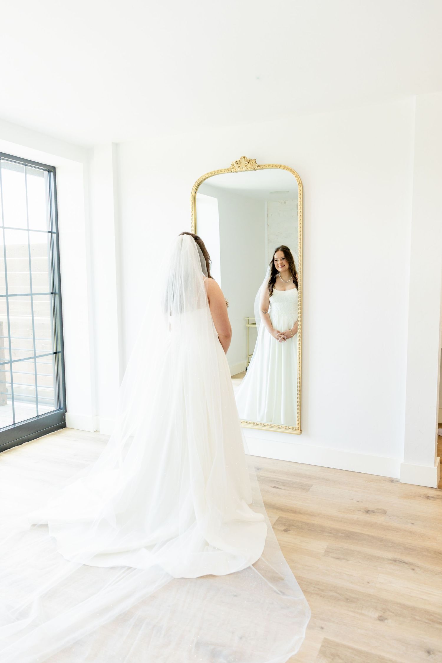 Bride in white gown and veil admires her reflection in an ornate gold floor mirror in a bright, airy bridal suite.