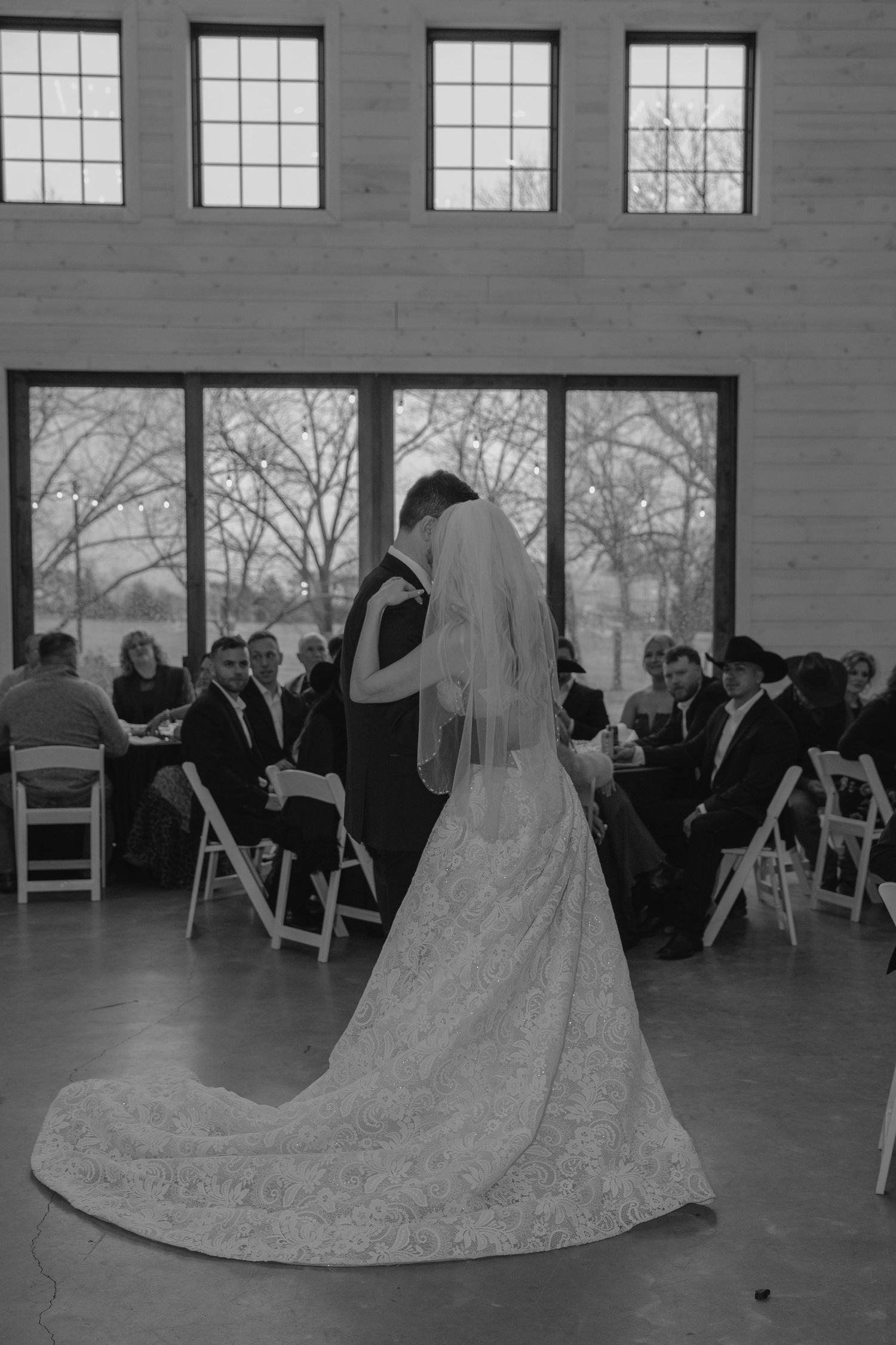 Black and white photo of bride and groom sharing first dance at indoor wedding ceremony with large windows.