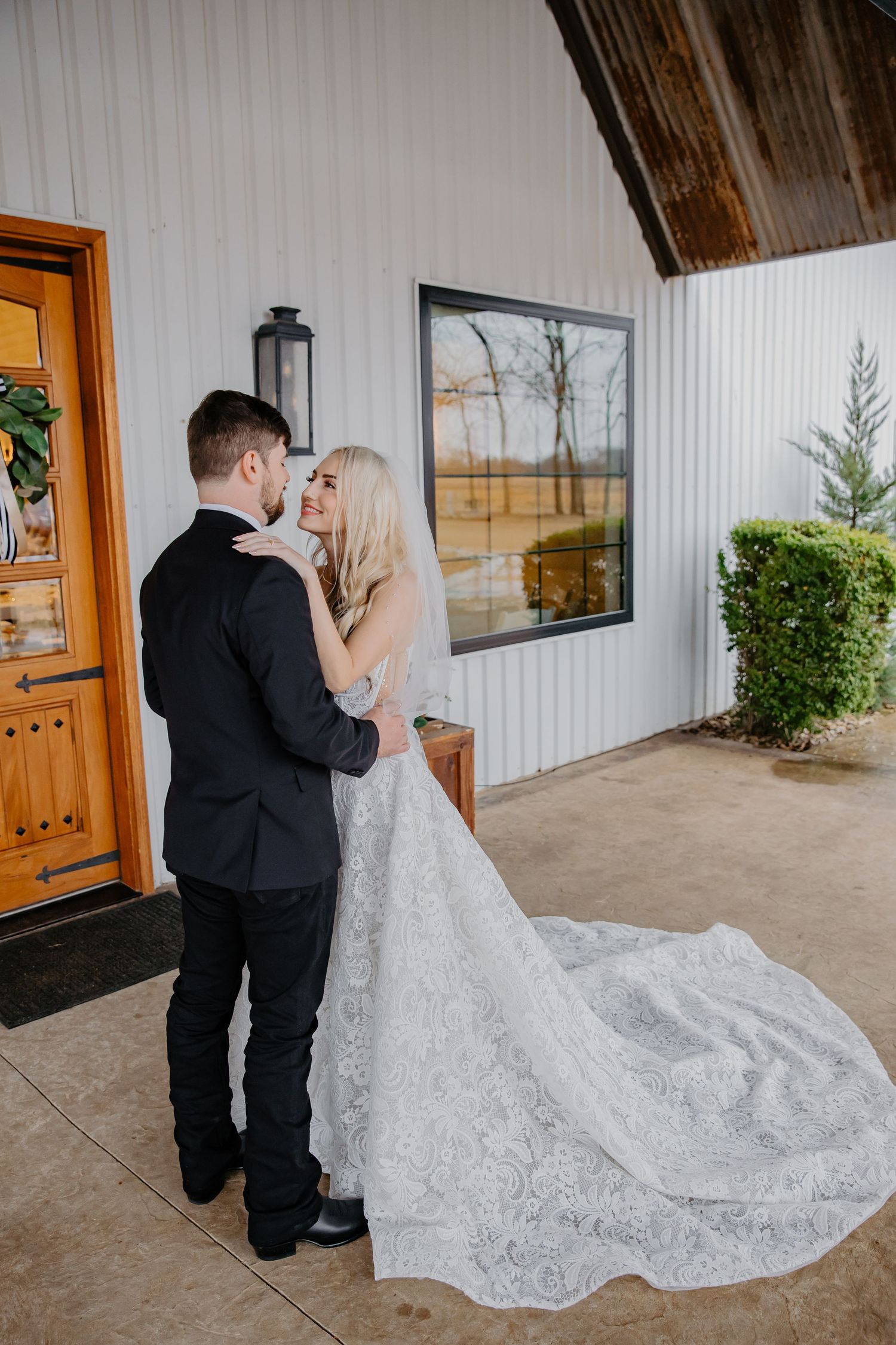 A bride in a lace gown with a long train embraces her groom in a black suit outside a white barn venue.