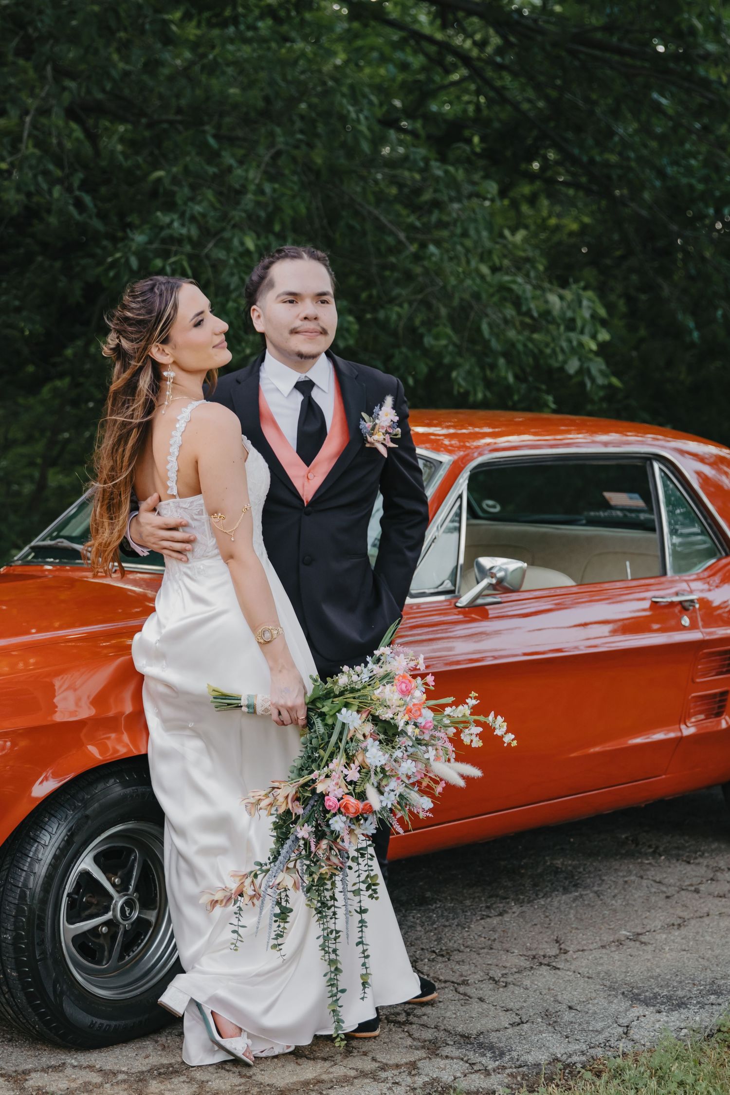 A wedding couple poses elegantly beside a classic orange car against a backdrop of green trees.