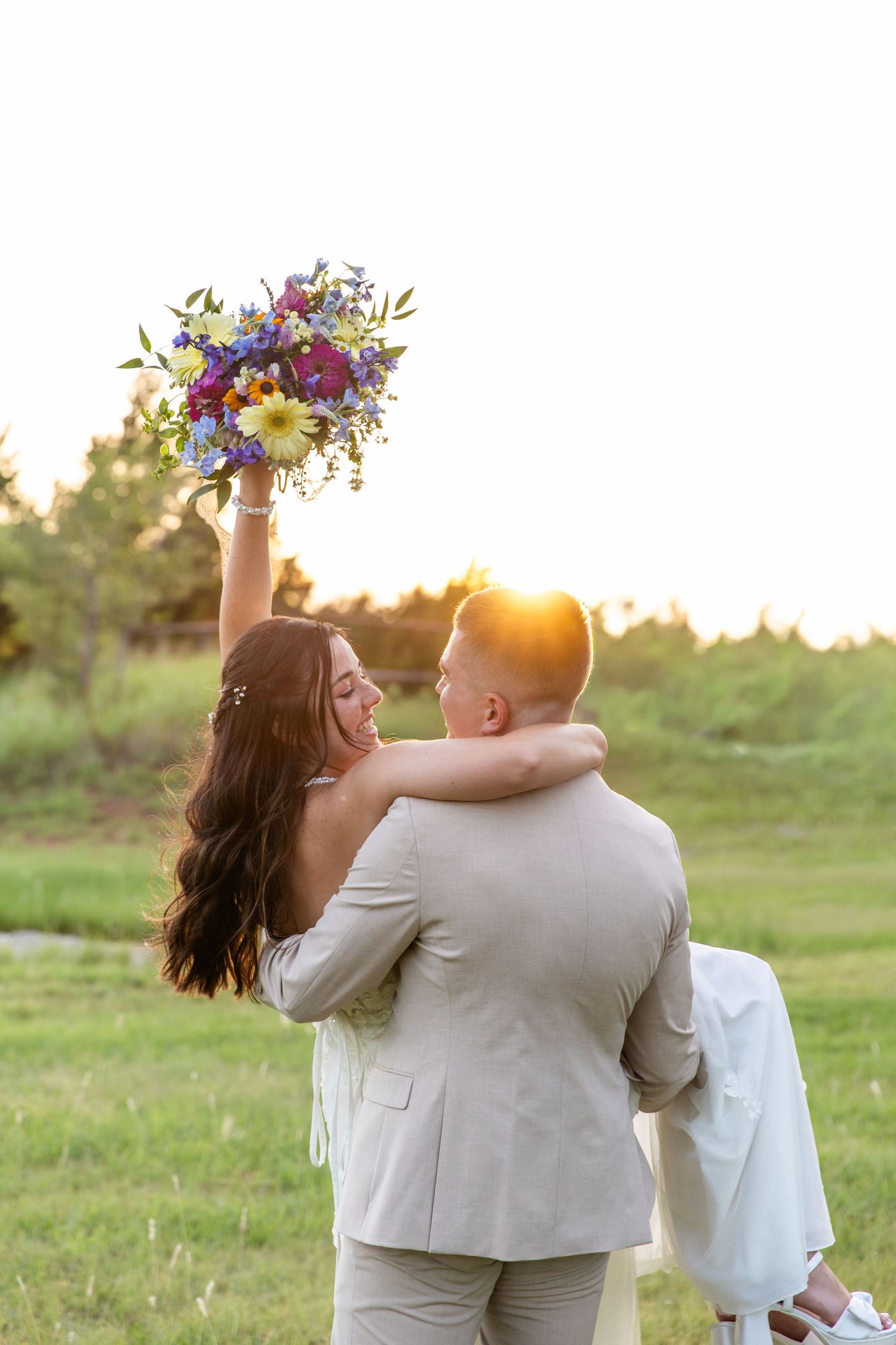 A joyful bride raises her colorful bouquet while embracing her groom at sunset in a lush green field.